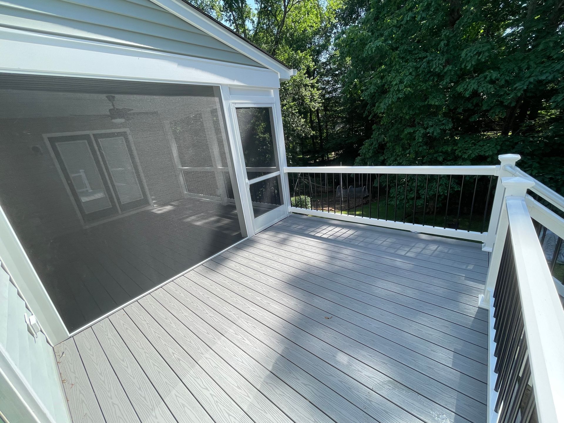 Sunlit white deck with railing beside a screened porch and green trees beyond
