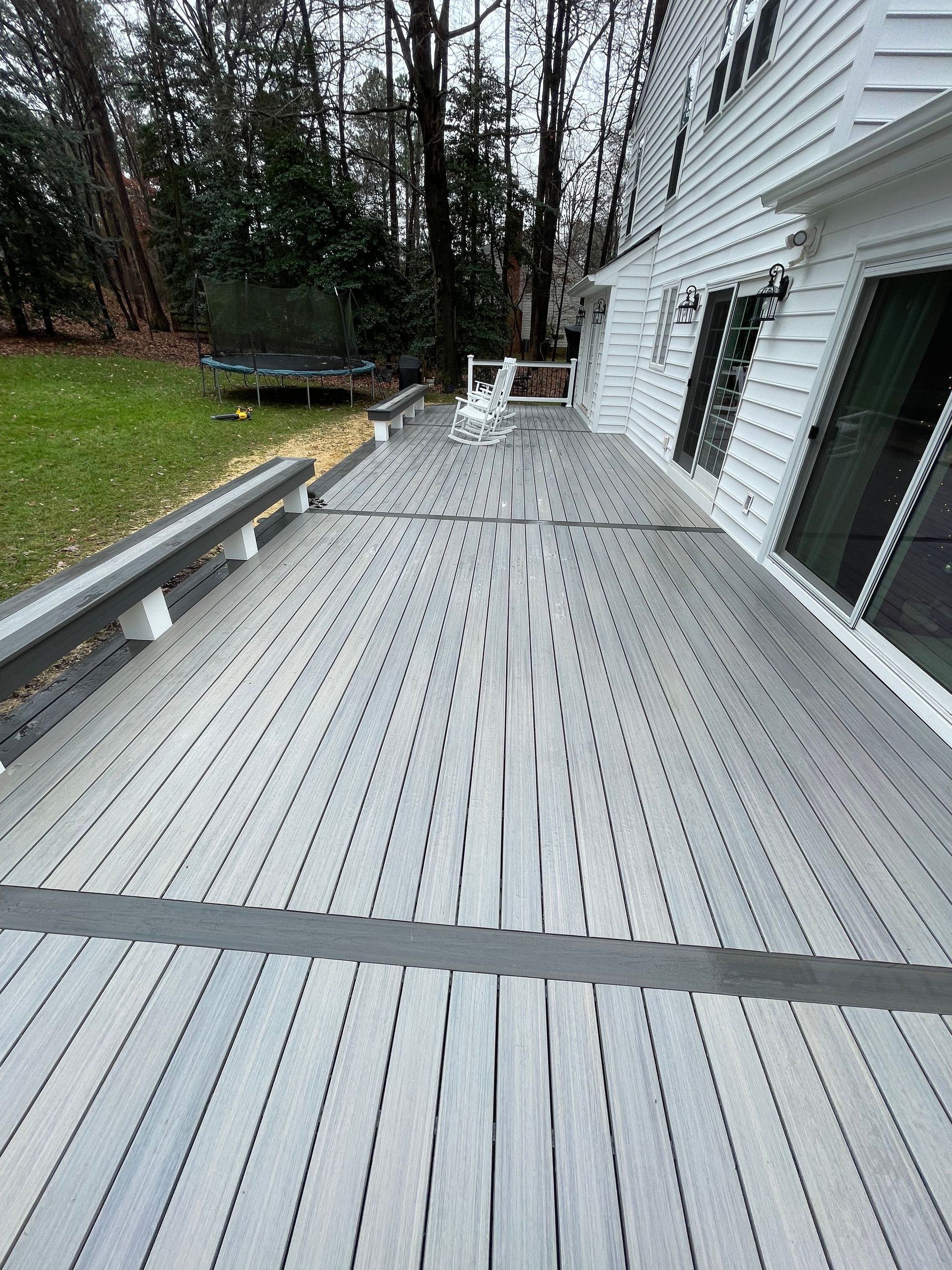 White wooden deck beside a house, bordered by railing and trees, with a chair at the far end.