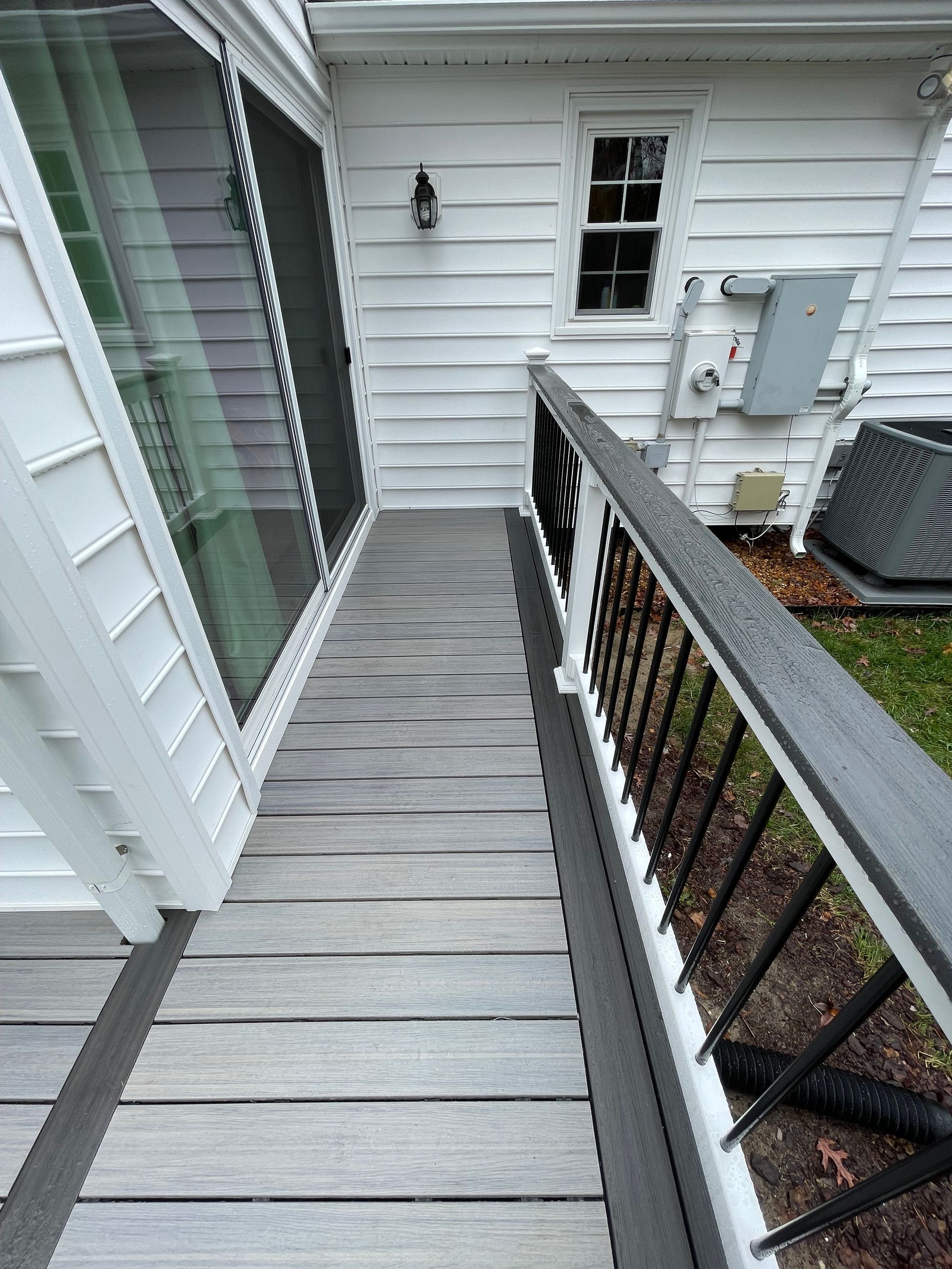 Narrow gray wooden walkway beside a white house, with black railing and utility meters ahead.