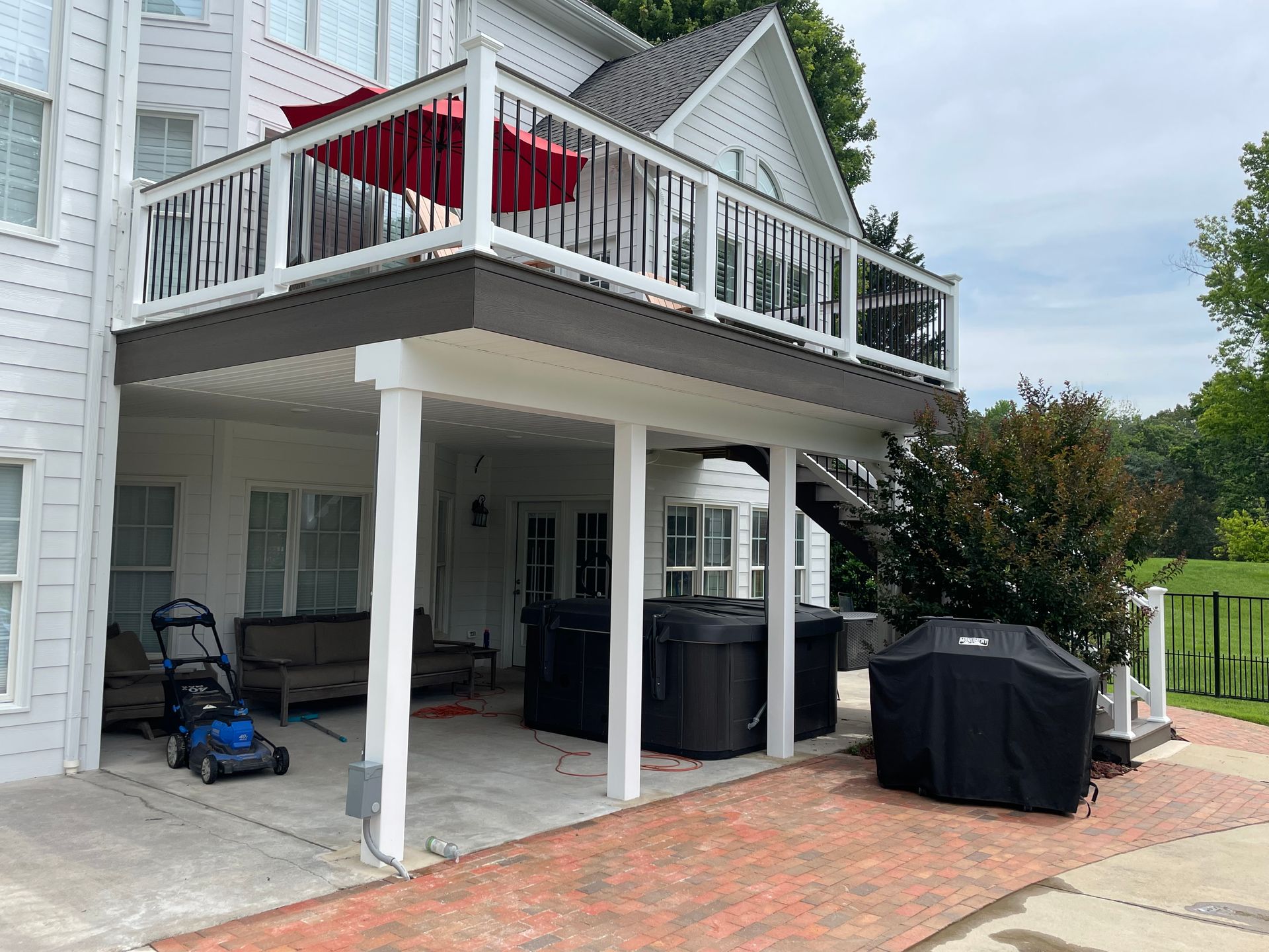 White house patio with second-story deck, red patio umbrella, hot tub, grill, and lawn equipment