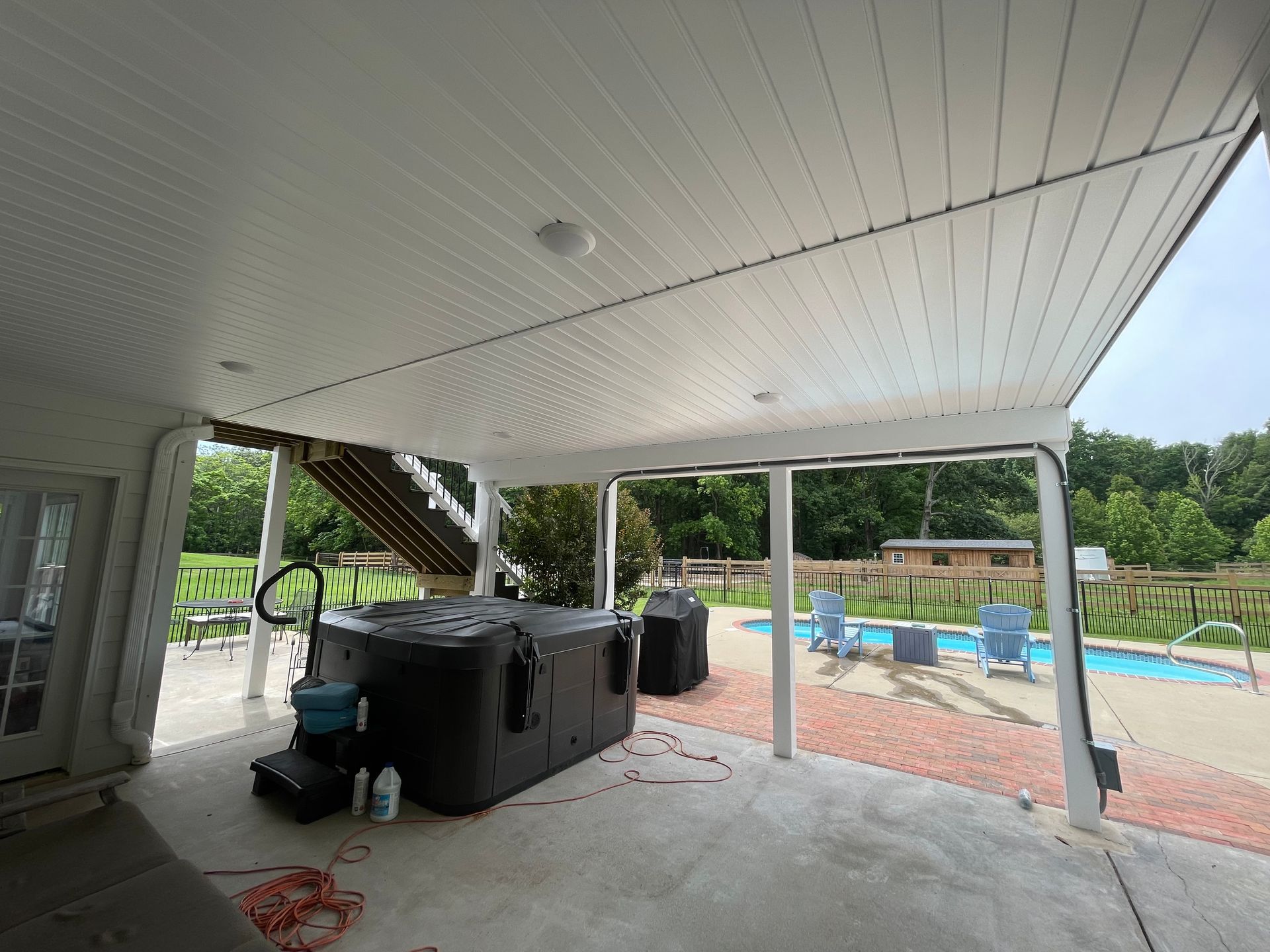 Covered patio beside a pool with hot tubs, lounge chairs, and a view of trees beyond the fence