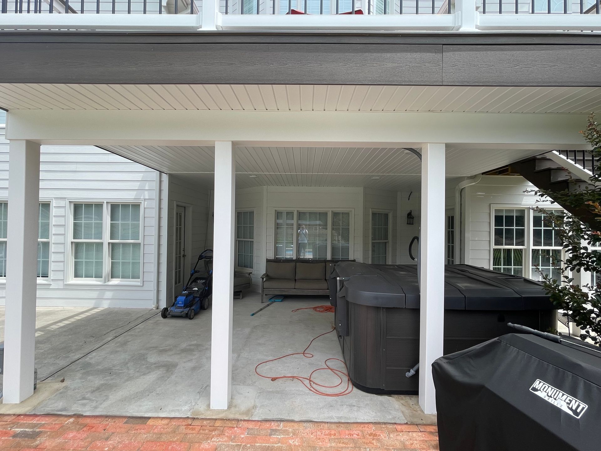 Covered patio with white pillars, gray bins, and a black grill under a raised deck.