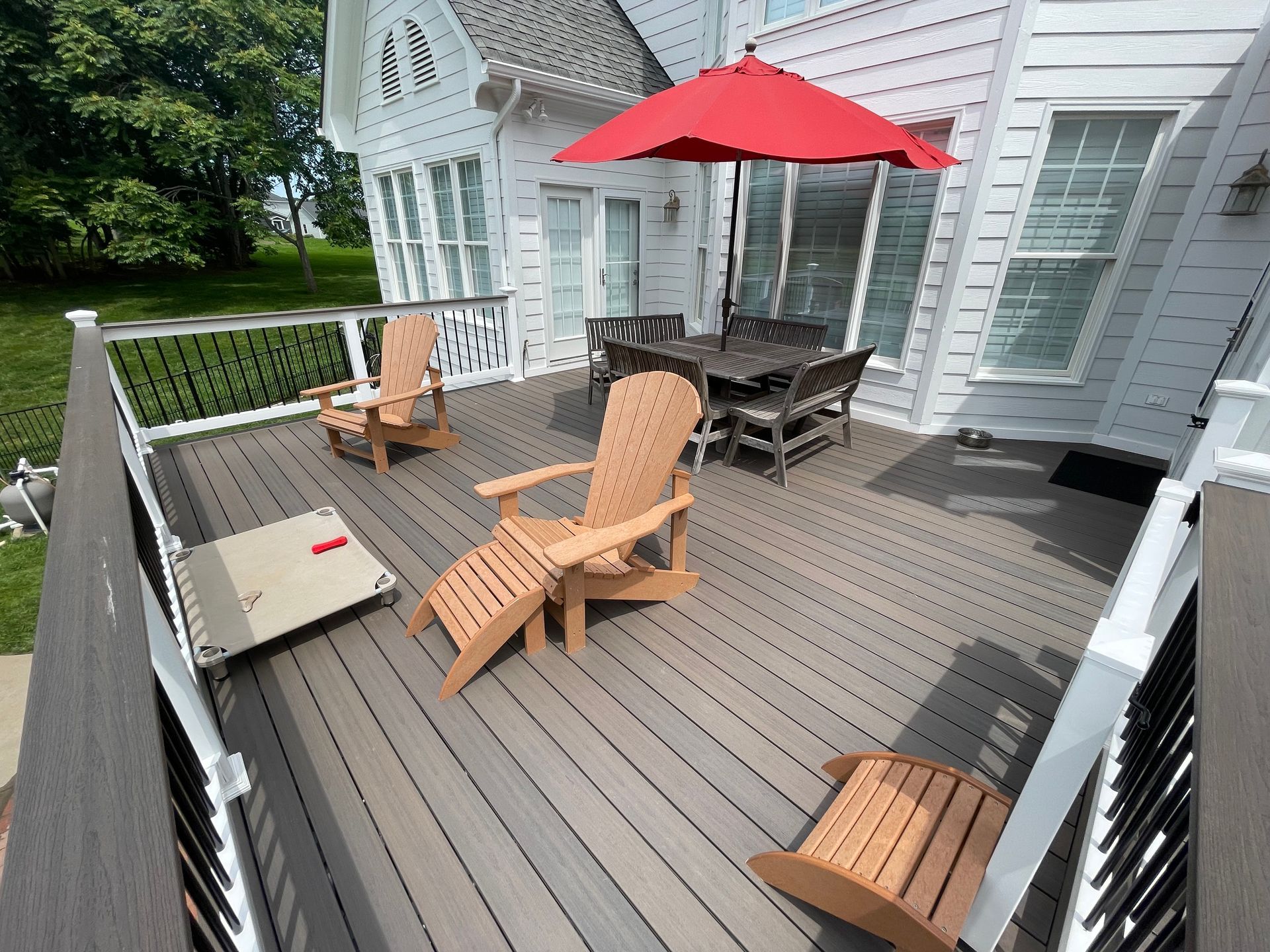 White house deck with Adirondack chairs, a red umbrella, and a cushioned bench overlooking a green yard