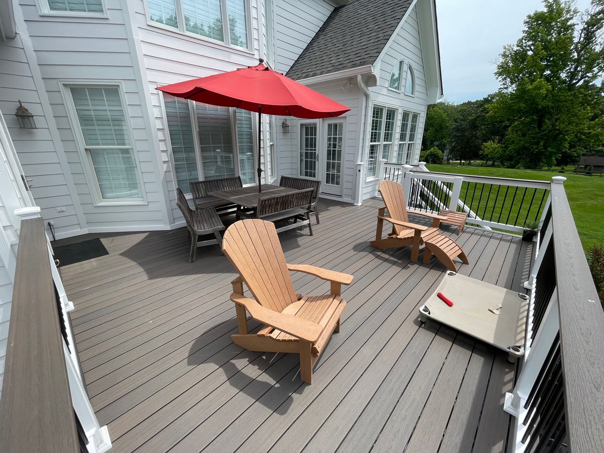 White house deck with red umbrella, wooden chairs, and a grill overlooking a grassy yard.