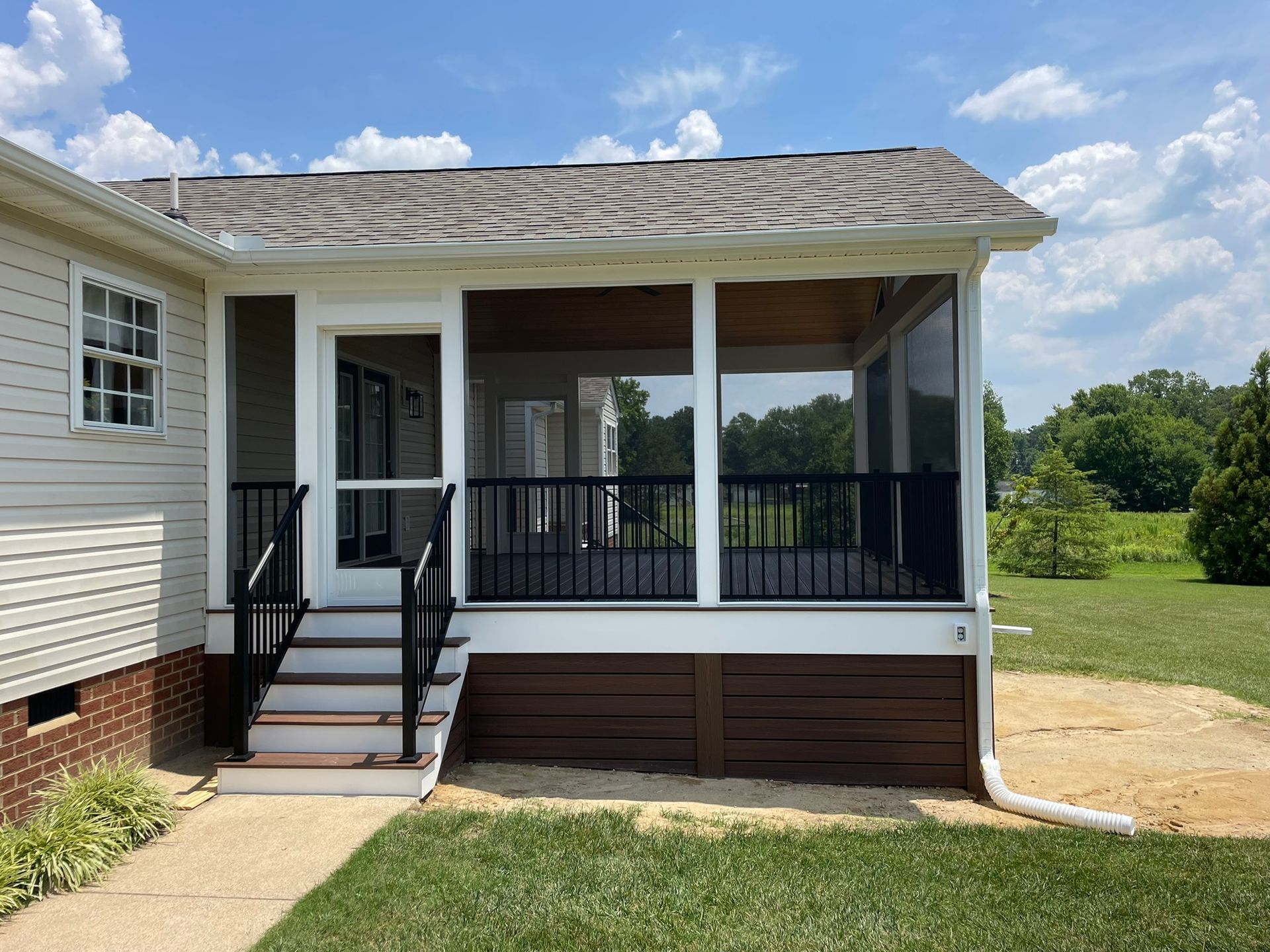 Covered porch with black railing and stairs on a light-sided house, next to an open grassy yard.