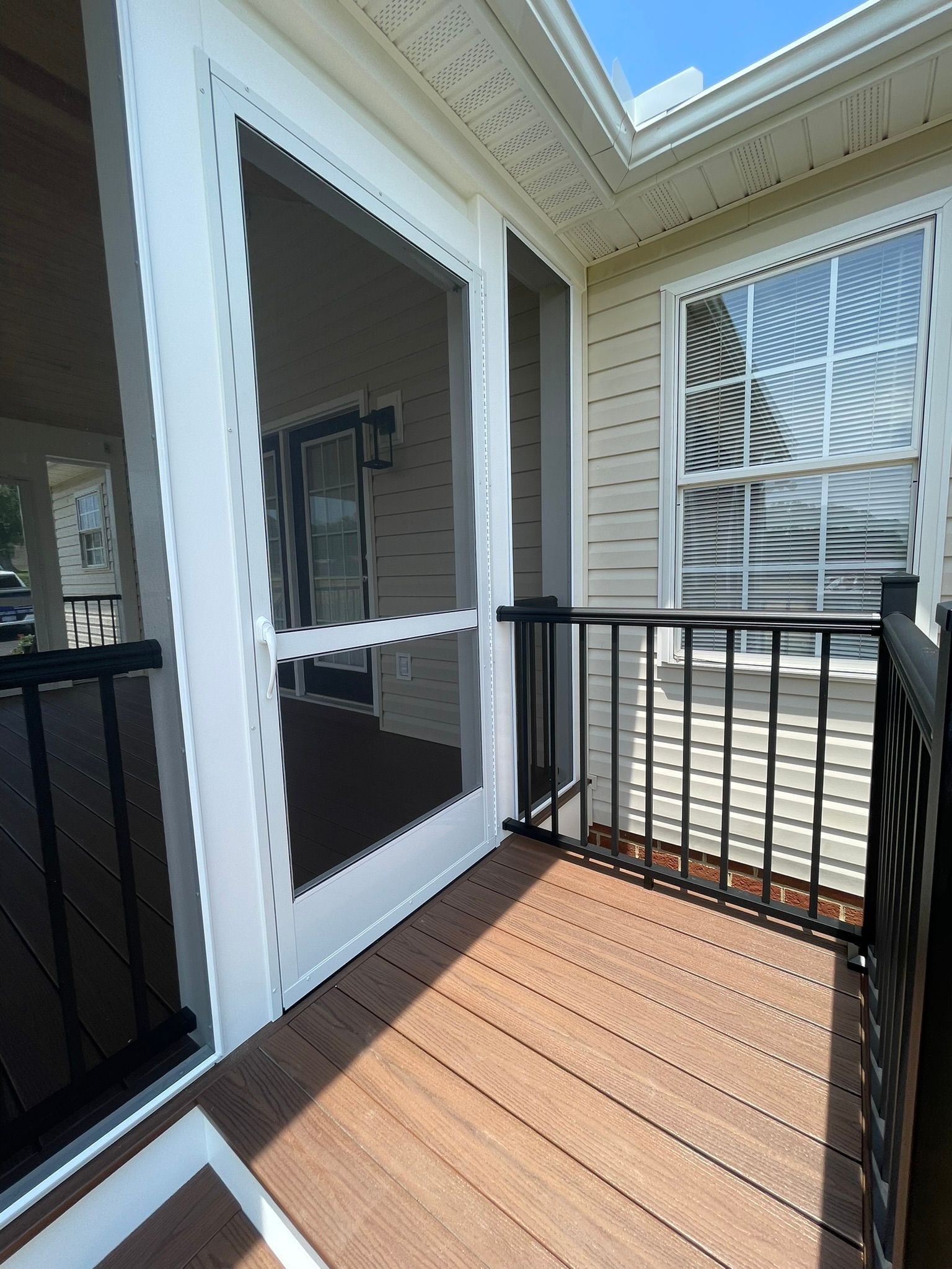 Small wooden balcony with black railing and white screened door beside a house window.
