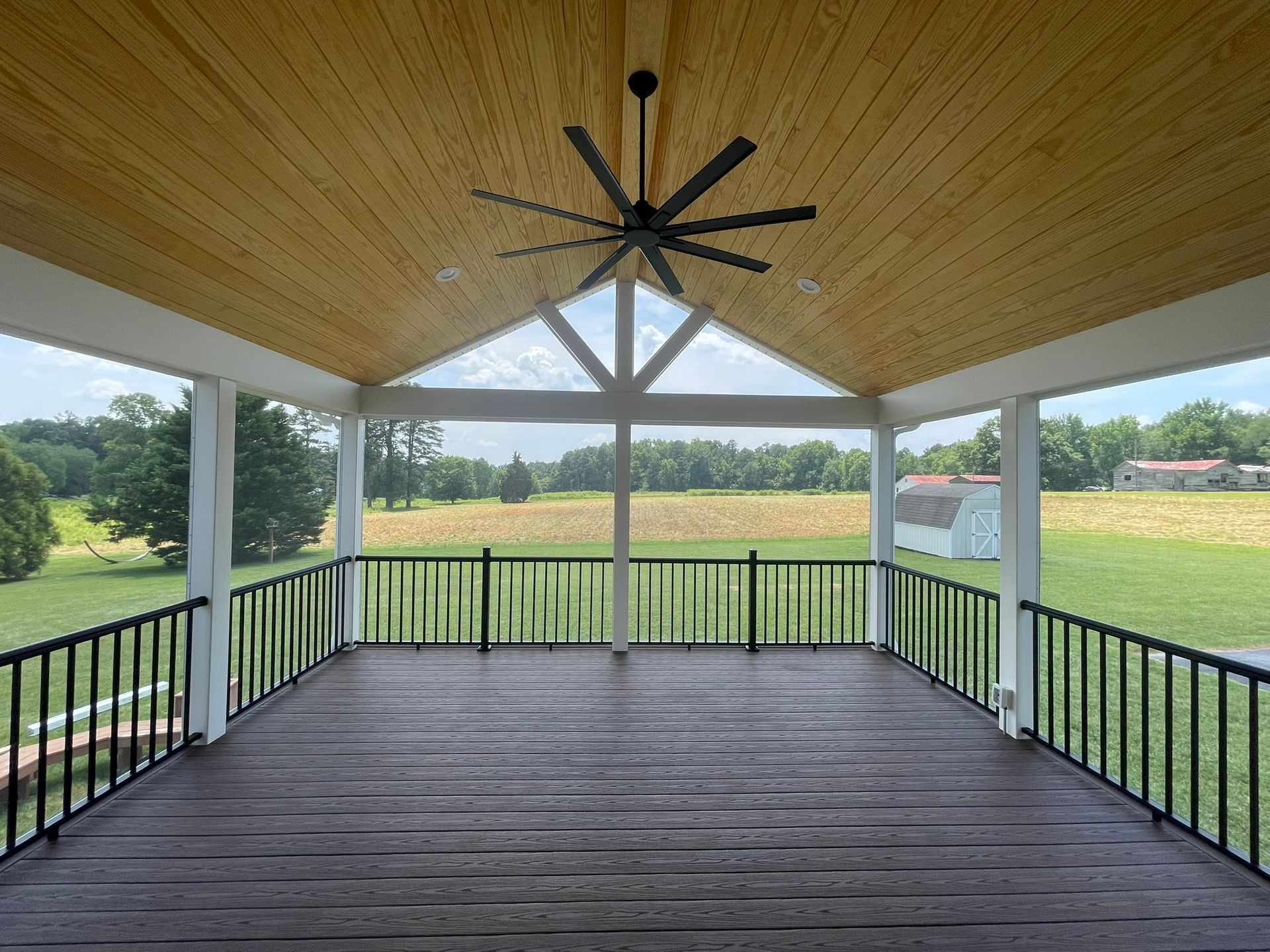 Covered wooden deck overlooking a green field, with black railings and a ceiling fan under a vaulted roof.
