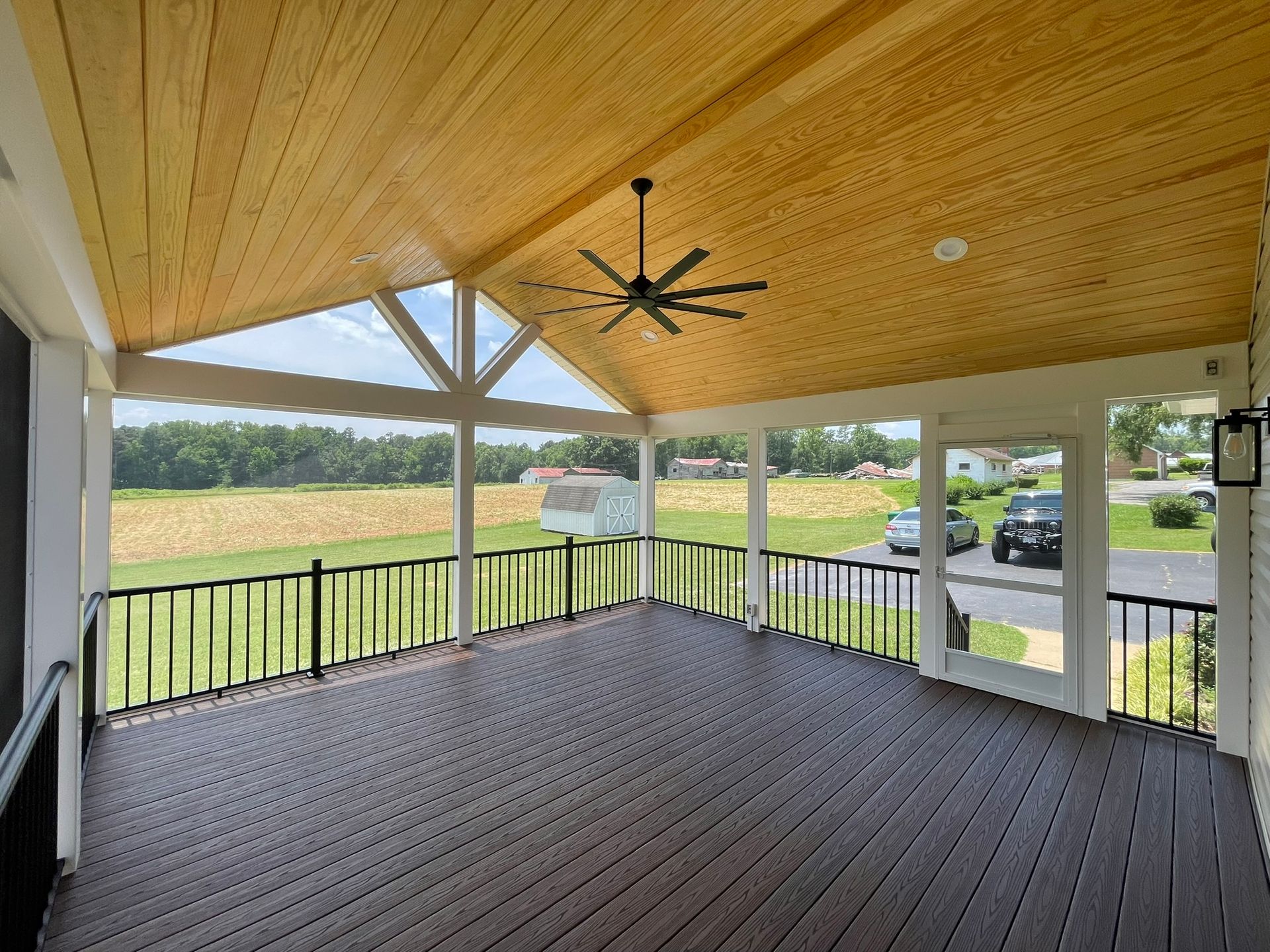 Covered porch with wood ceiling, black railing, and open view of a grassy backyard and parked cars.