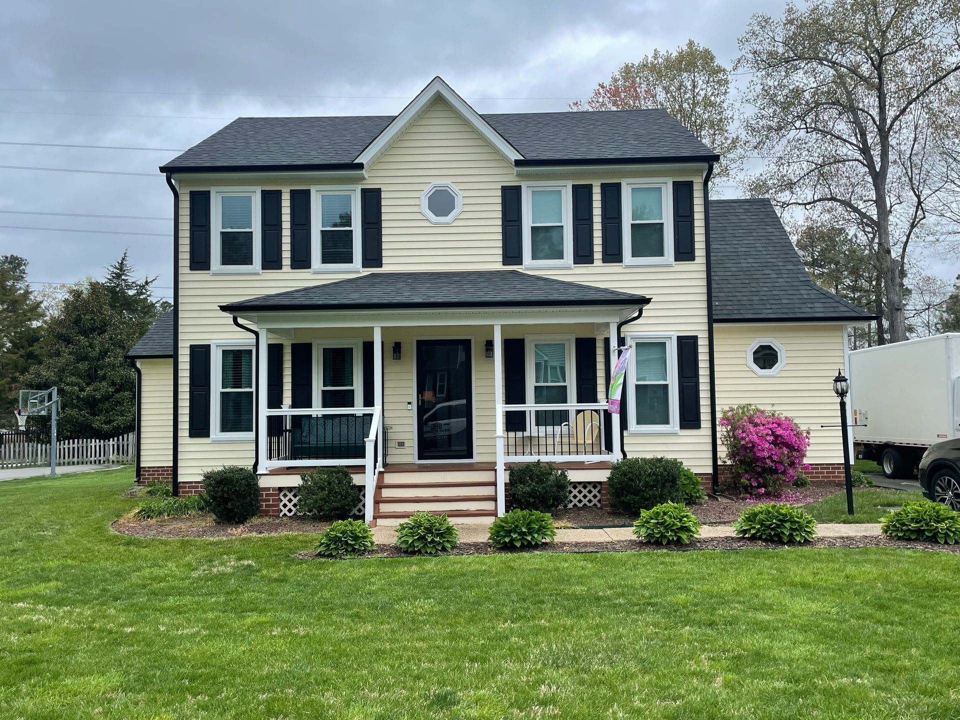 Two-story yellow house with black shutters, front porch, and a large green lawn under an overcast sky