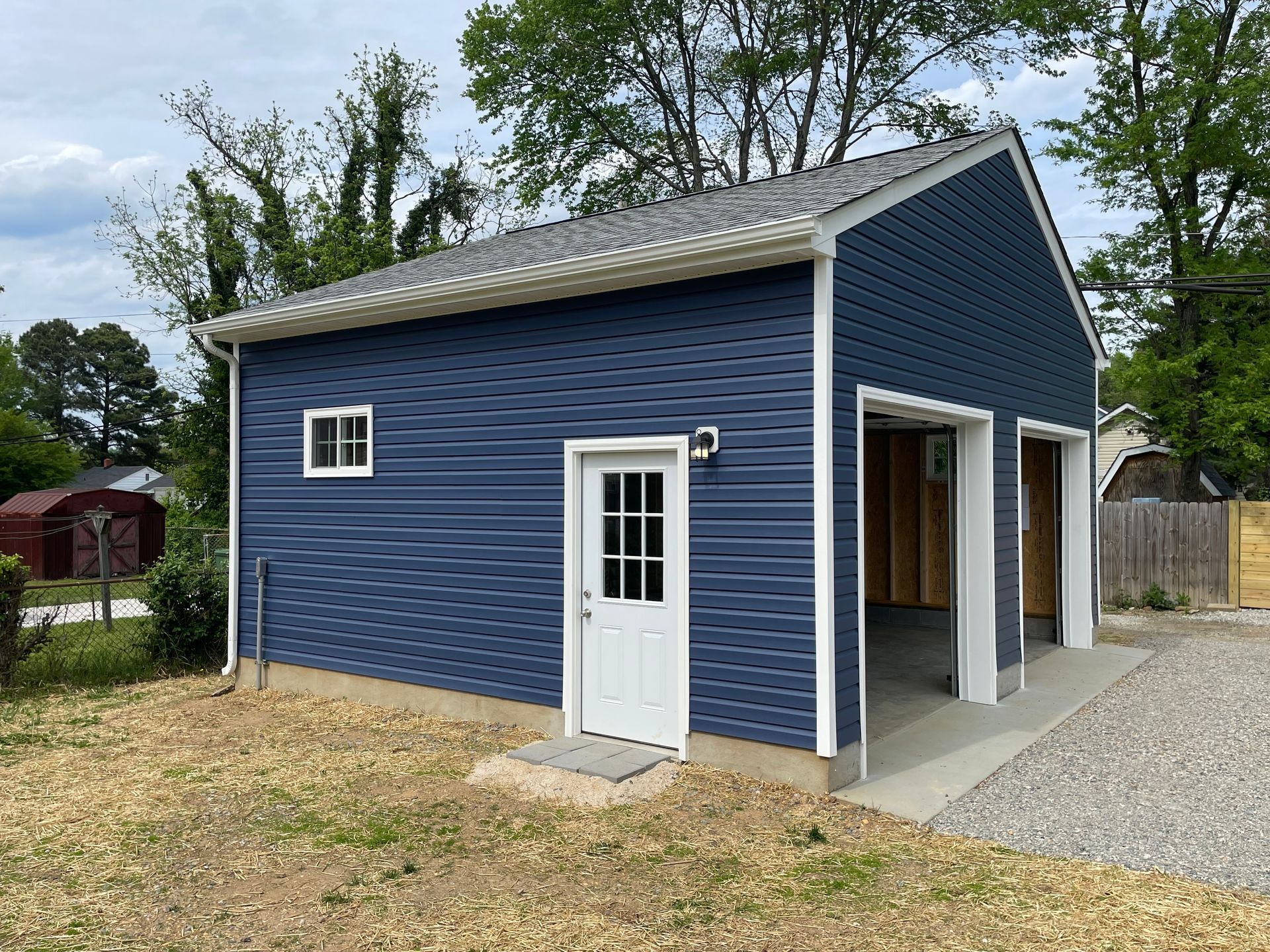Blue detached garage with white trim, side door, and open bays beside a gravel driveway