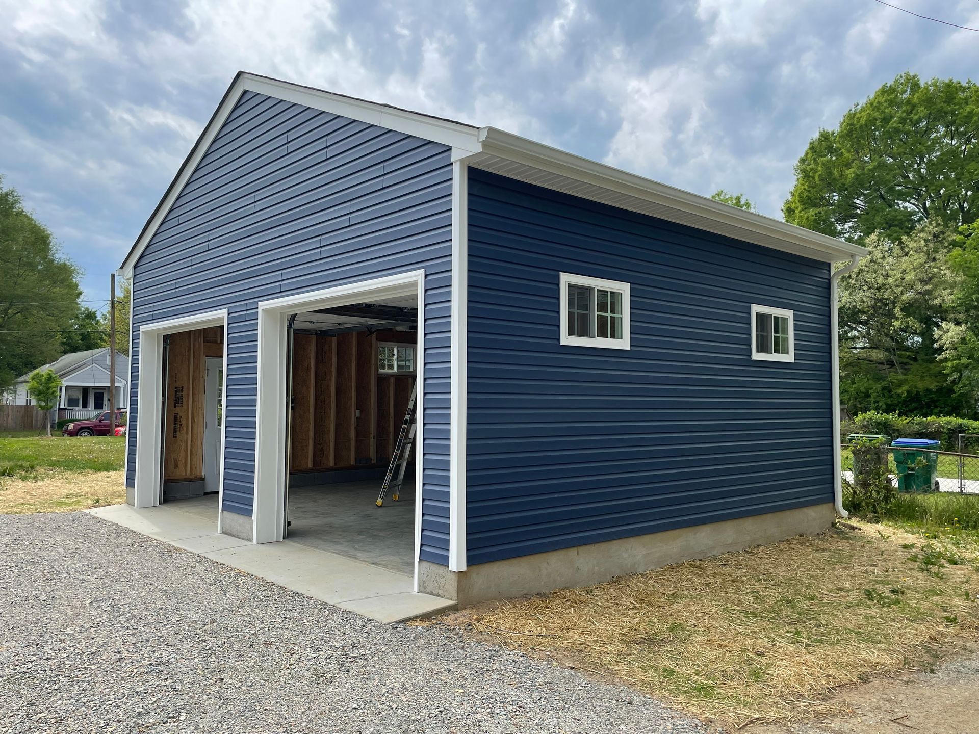Blue detached garage with white trim and two open bays on a gravel driveway under a cloudy sky