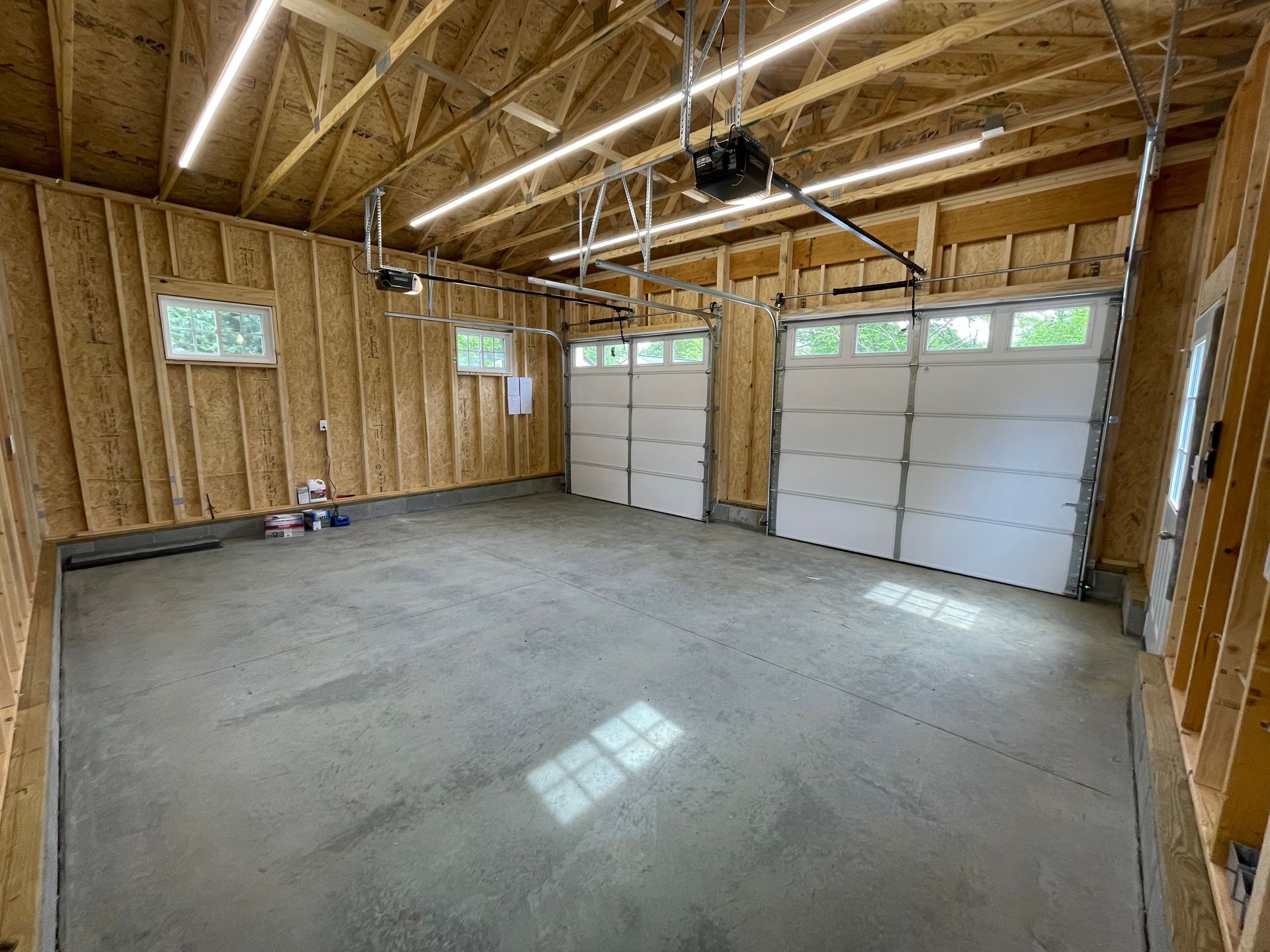 Empty unfinished garage with concrete floor, exposed wood framing, and two white garage doors.
