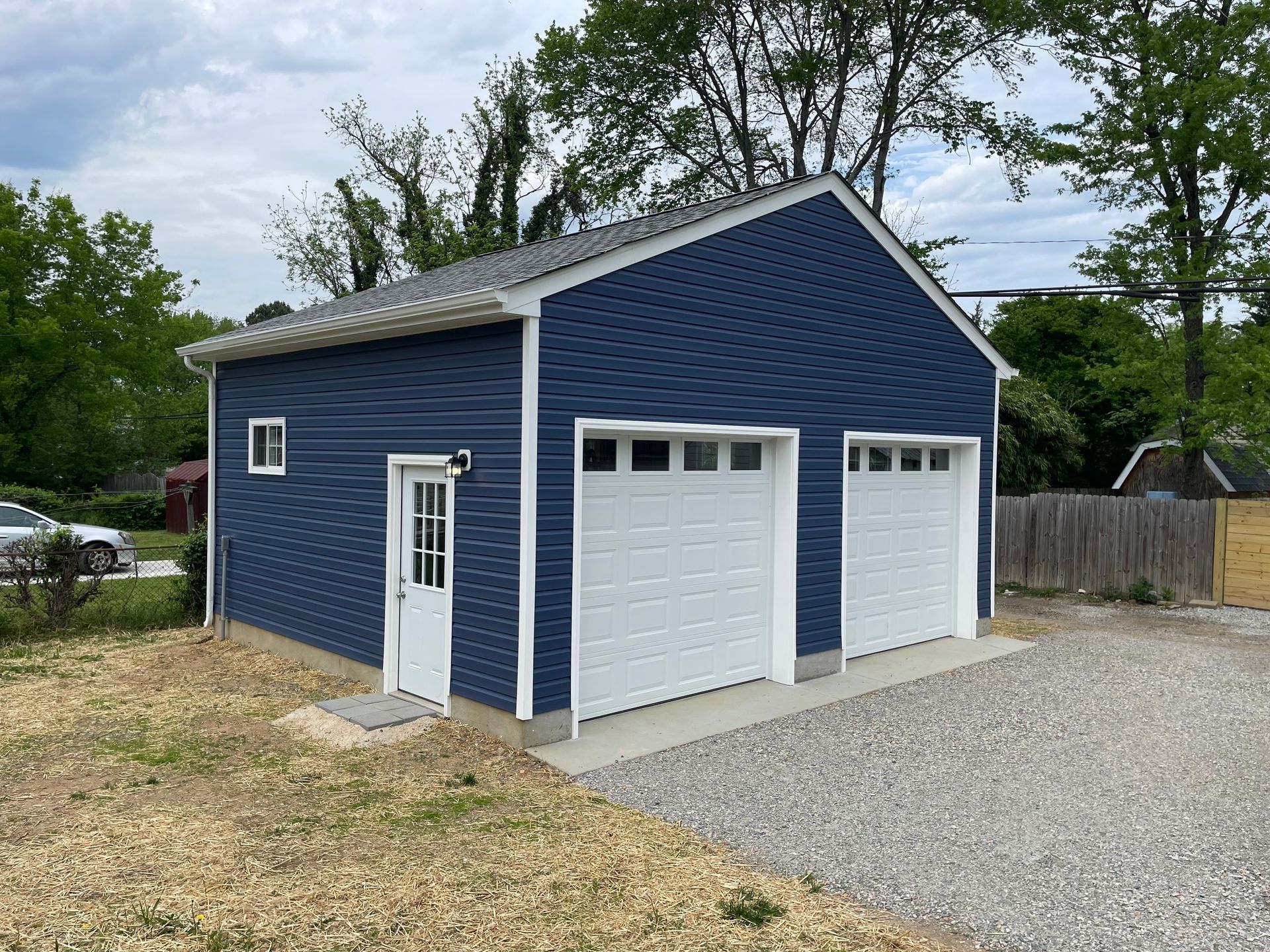 Blue detached garage with white doors and gravel driveway in a wooded yard