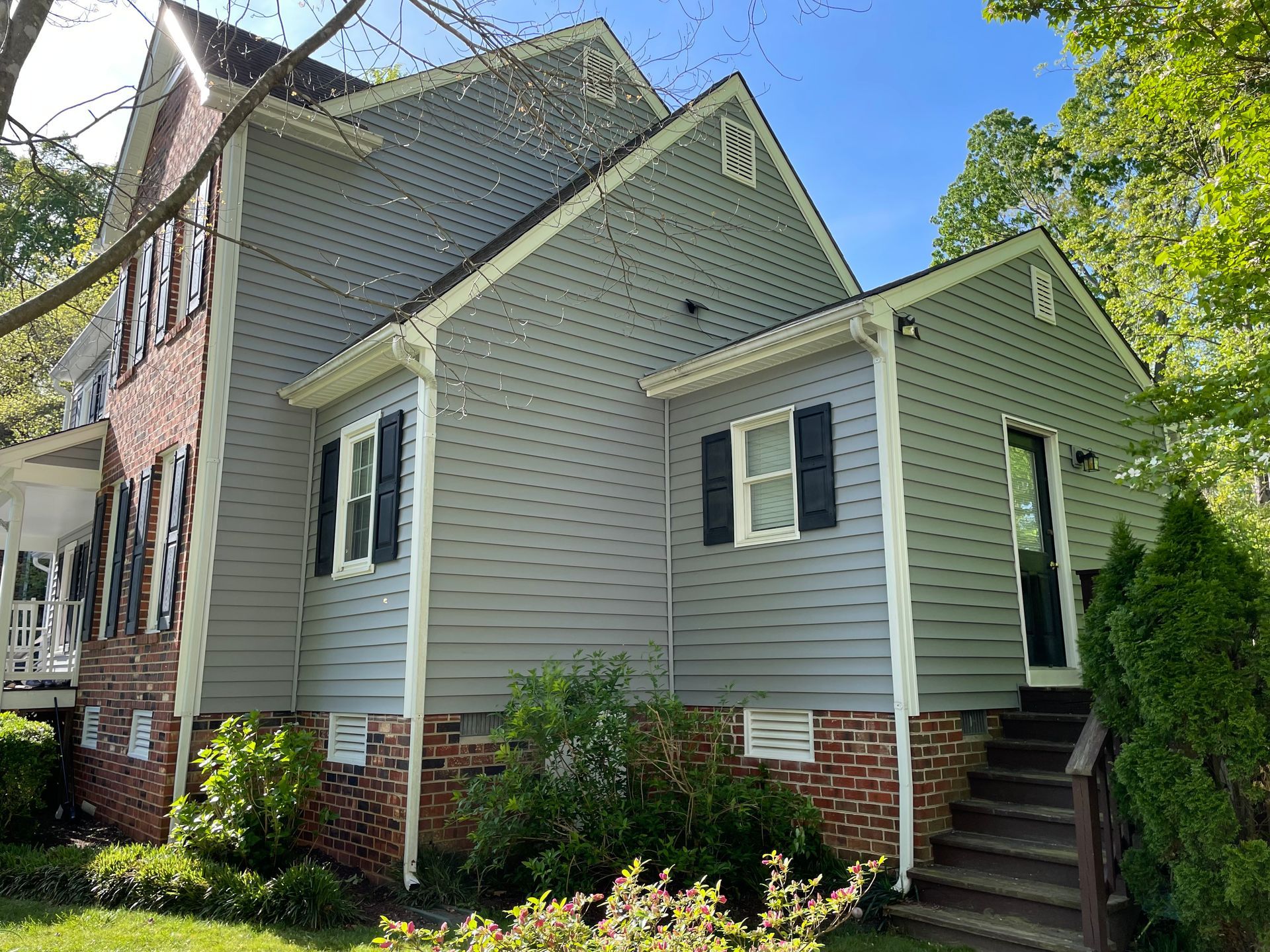 Gray house with white trim and brick foundation, viewed from the side with front steps and trees nearby