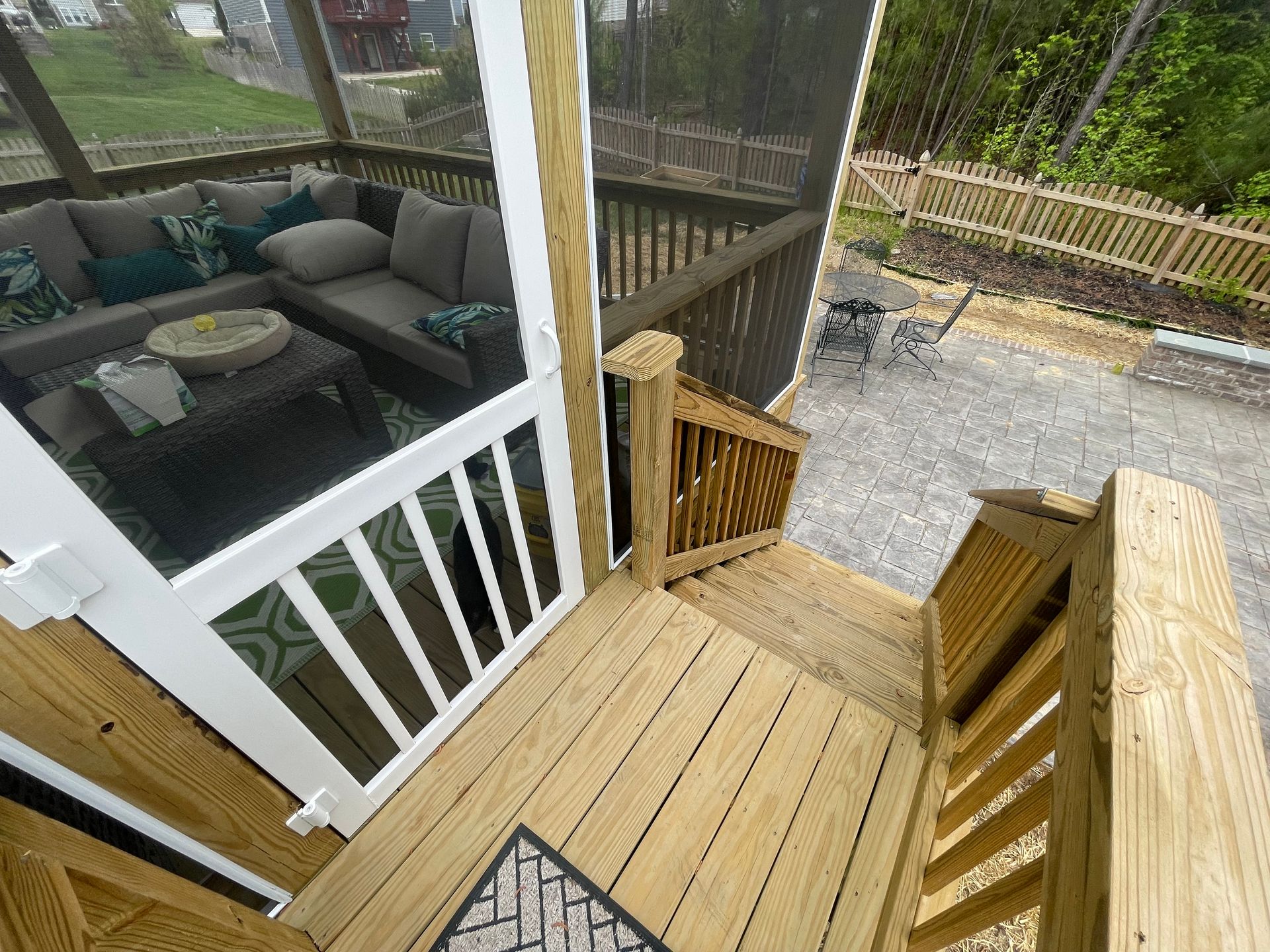 Wooden deck stairs beside a screened porch with patio seating and a gravel yard beyond