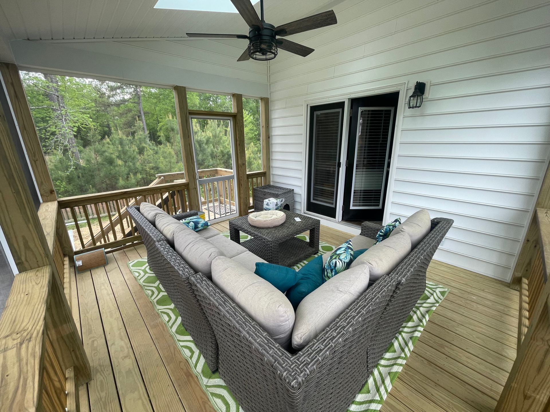 Covered porch with gray sectional sofa, fire pit table, ceiling fan, and wooden railing overlooking trees