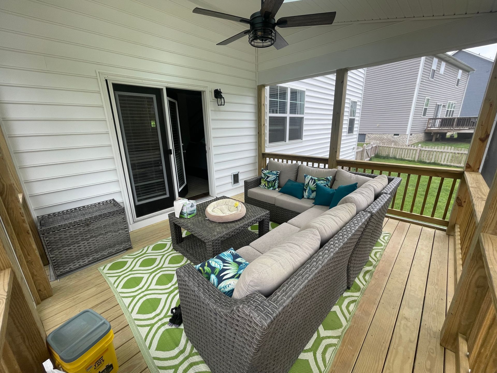 Covered porch with gray sectional sofa, green rug, and railing overlooking a yard and neighboring houses