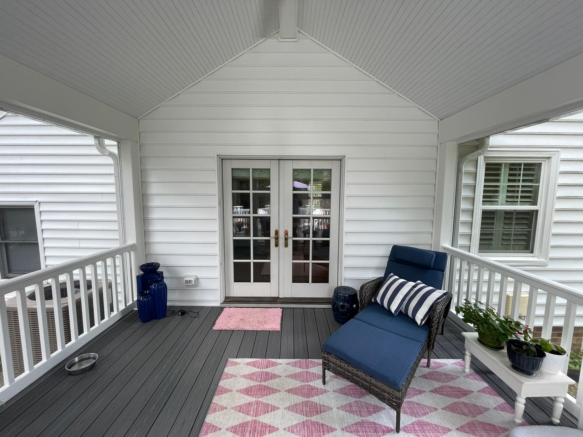 Covered porch with white siding, French doors, blue chair, pink rugs, and potted plants.