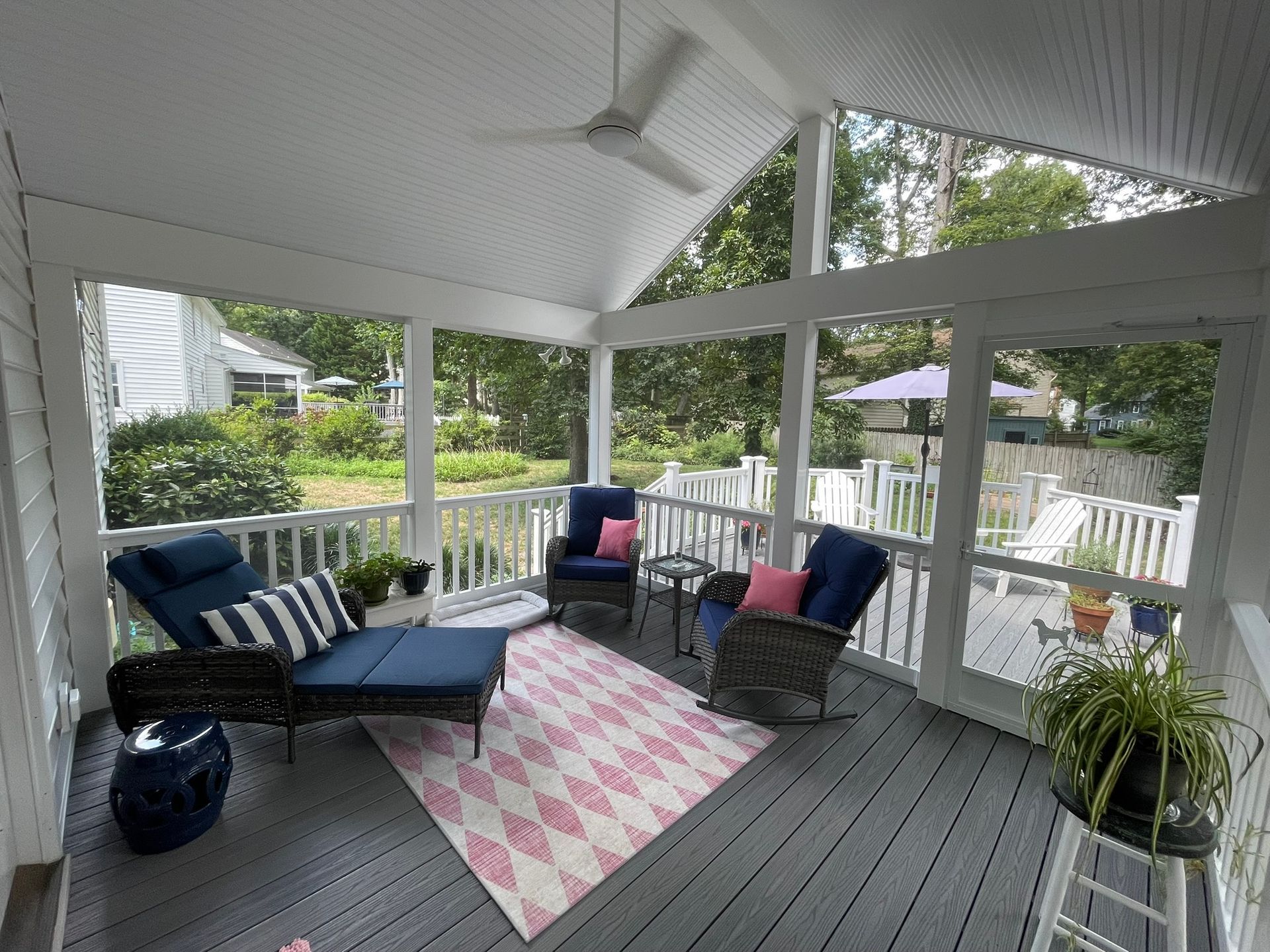 Screened porch with gray deck, wicker chairs, blue chaise, pink rug, and potted plants.