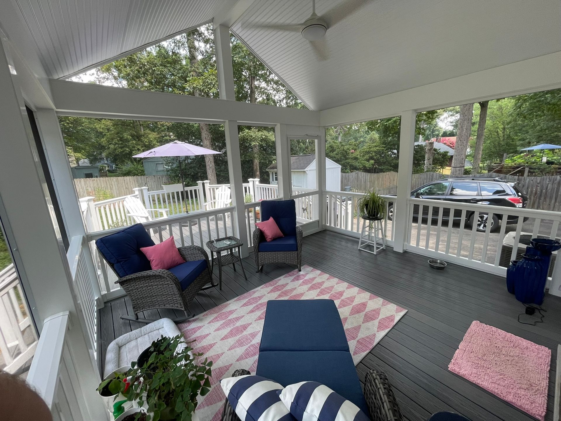 Covered porch with wicker seating, blue ottoman, pink rug, and white railings overlooking a street.