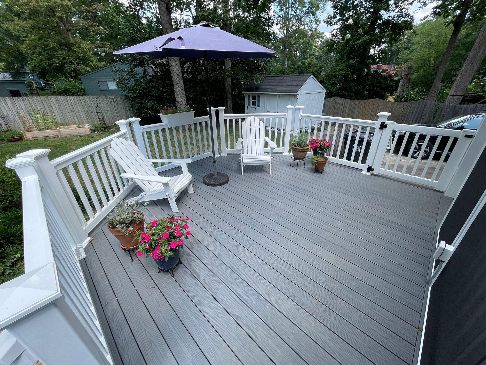 White deck with railing, chairs, umbrella, and potted flowers in a backyard.