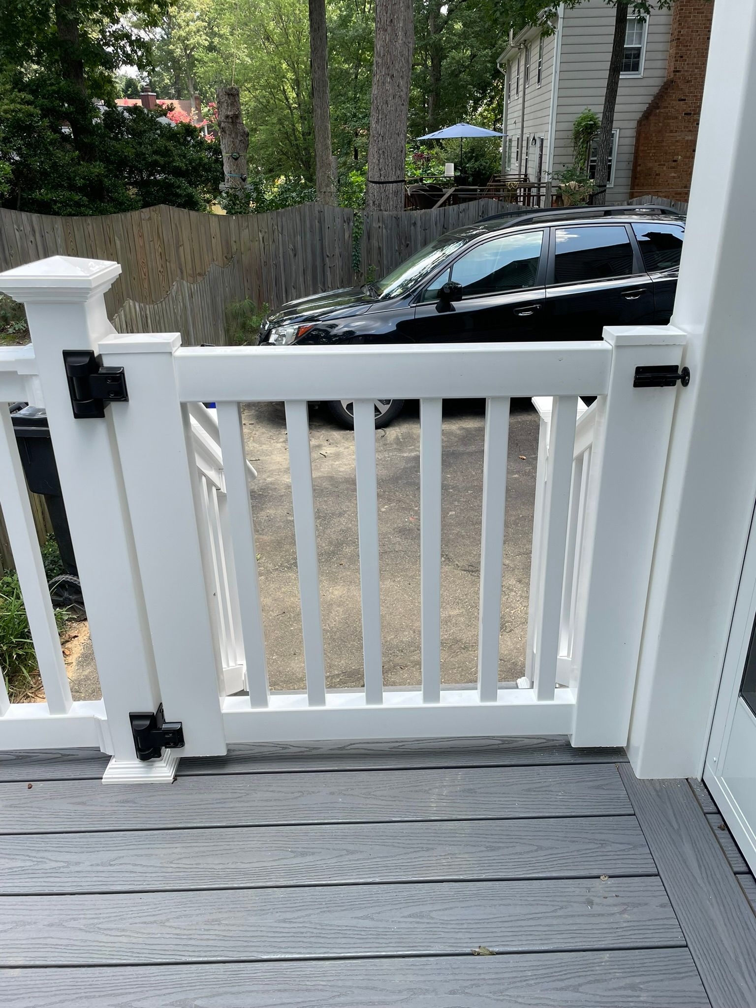 White porch gate with black latches, opening onto a driveway with a parked black car beyond.