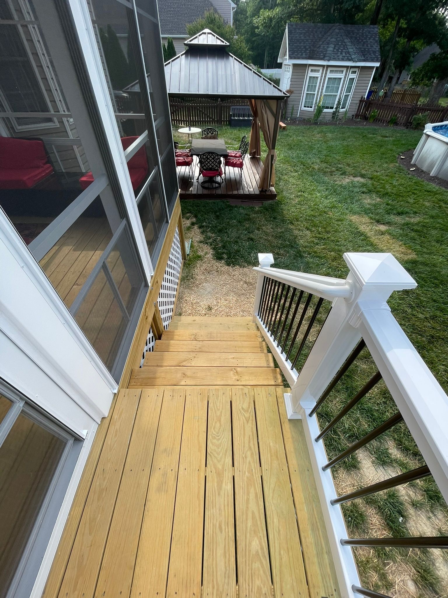 Wooden deck stairs leading to a backyard gazebo and green lawn, viewed from a porch.
