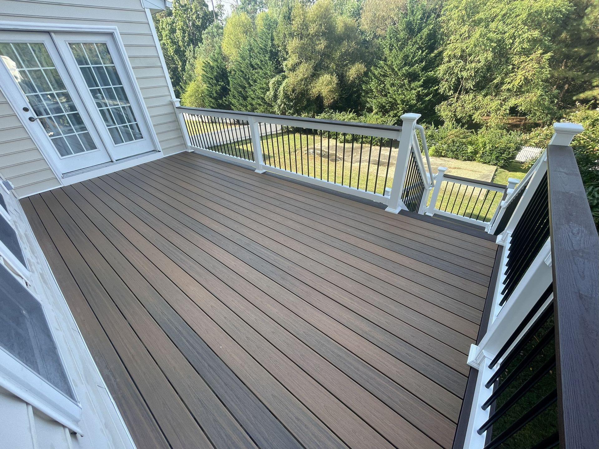 Spacious wooden deck with white railing beside a house, overlooking trees and a yard.