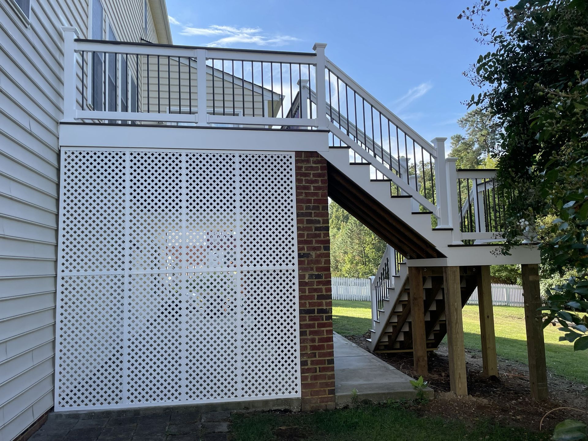 Raised wooden deck with white railing and staircase beside a house, with lattice skirting and brick supports