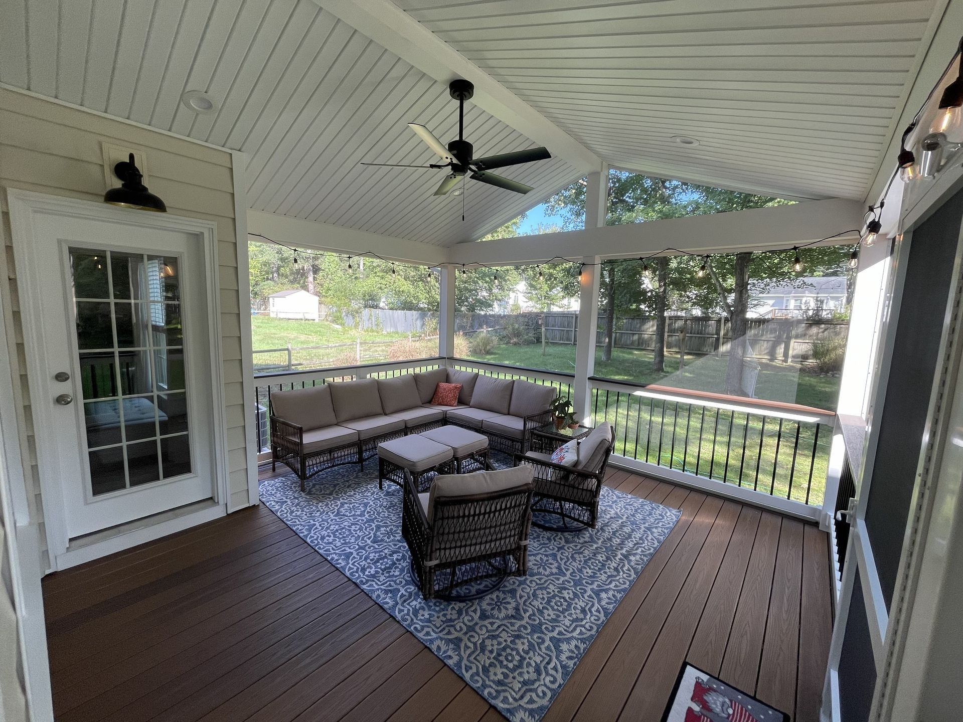 Covered porch with wicker seating, patterned rug, and ceiling fan overlooking a wooded yard