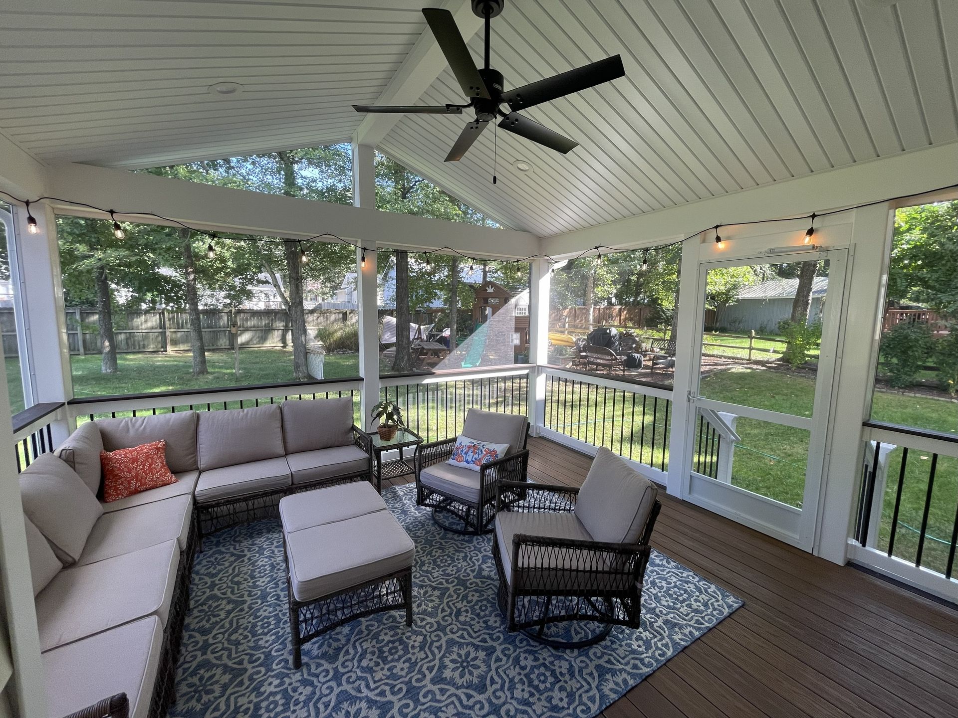 Covered screened porch with gray sectional, wicker chairs, coffee table, rug, and ceiling fan overlooking a yard