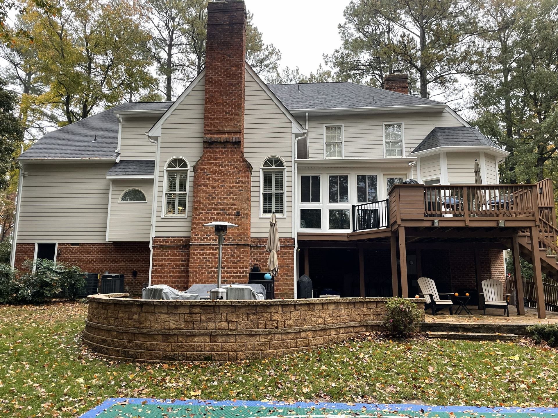 Back view of a beige house with a brick chimney, raised deck, and stone retaining wall in autumn