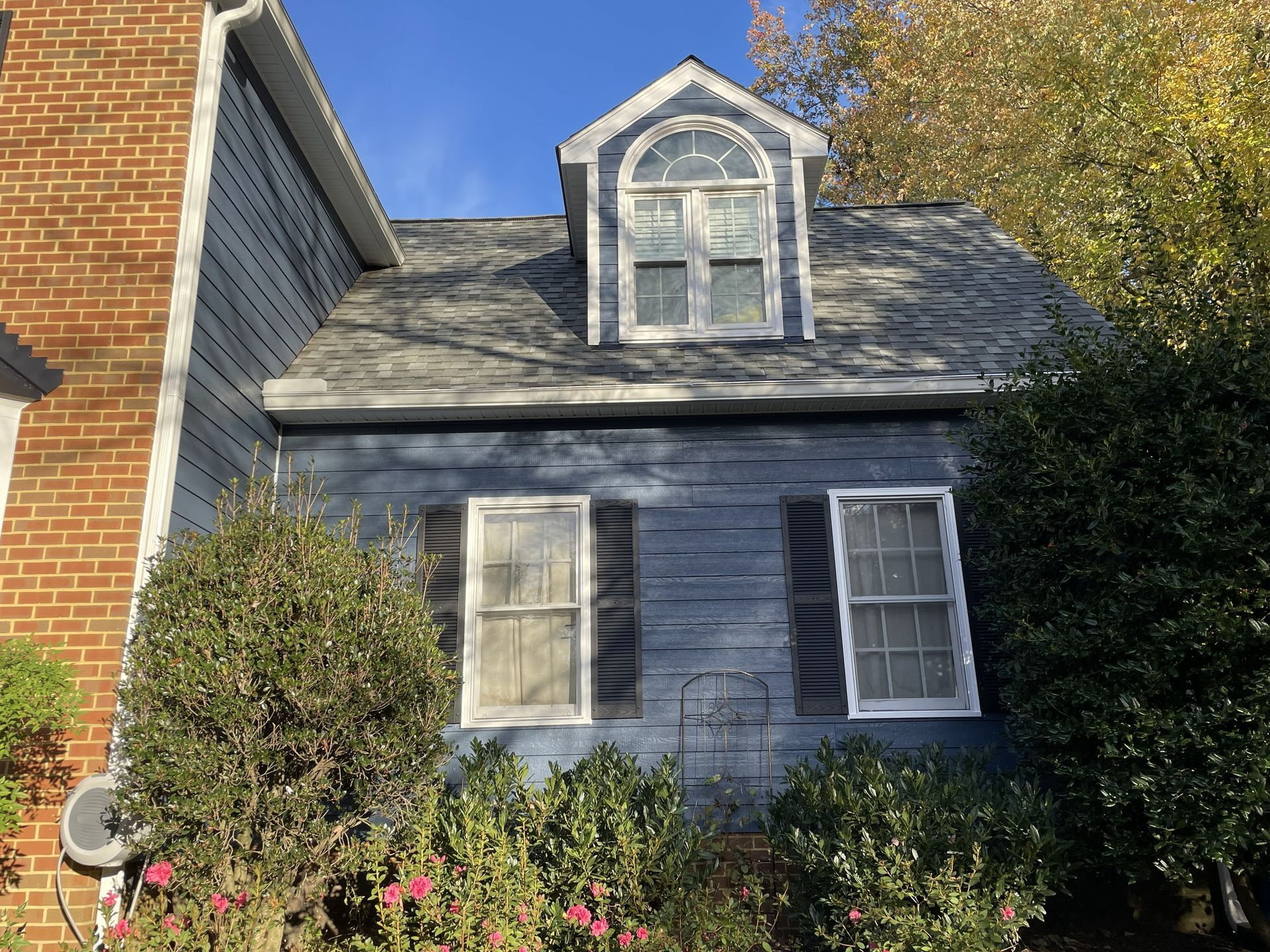 Blue house exterior with dormer windows, gray roof, and bushes in front under a clear sky