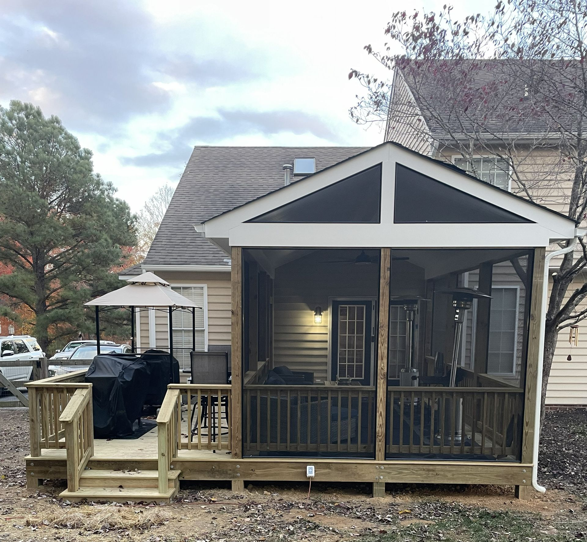 Wooden screened porch with black railing and steps attached to a beige house in a backyard.