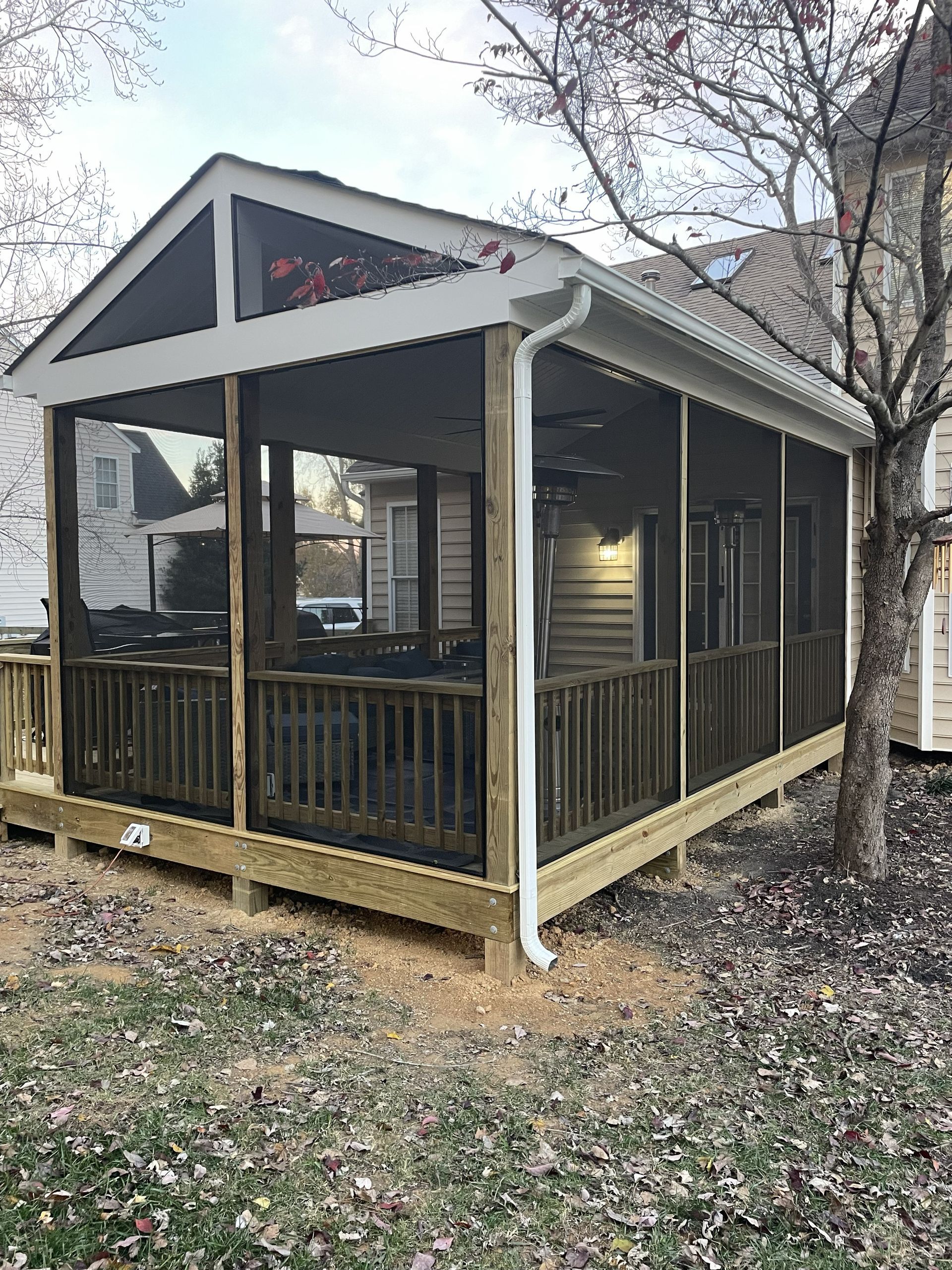 Small screened porch with wooden railing and gray roof beside a tree in a yard.