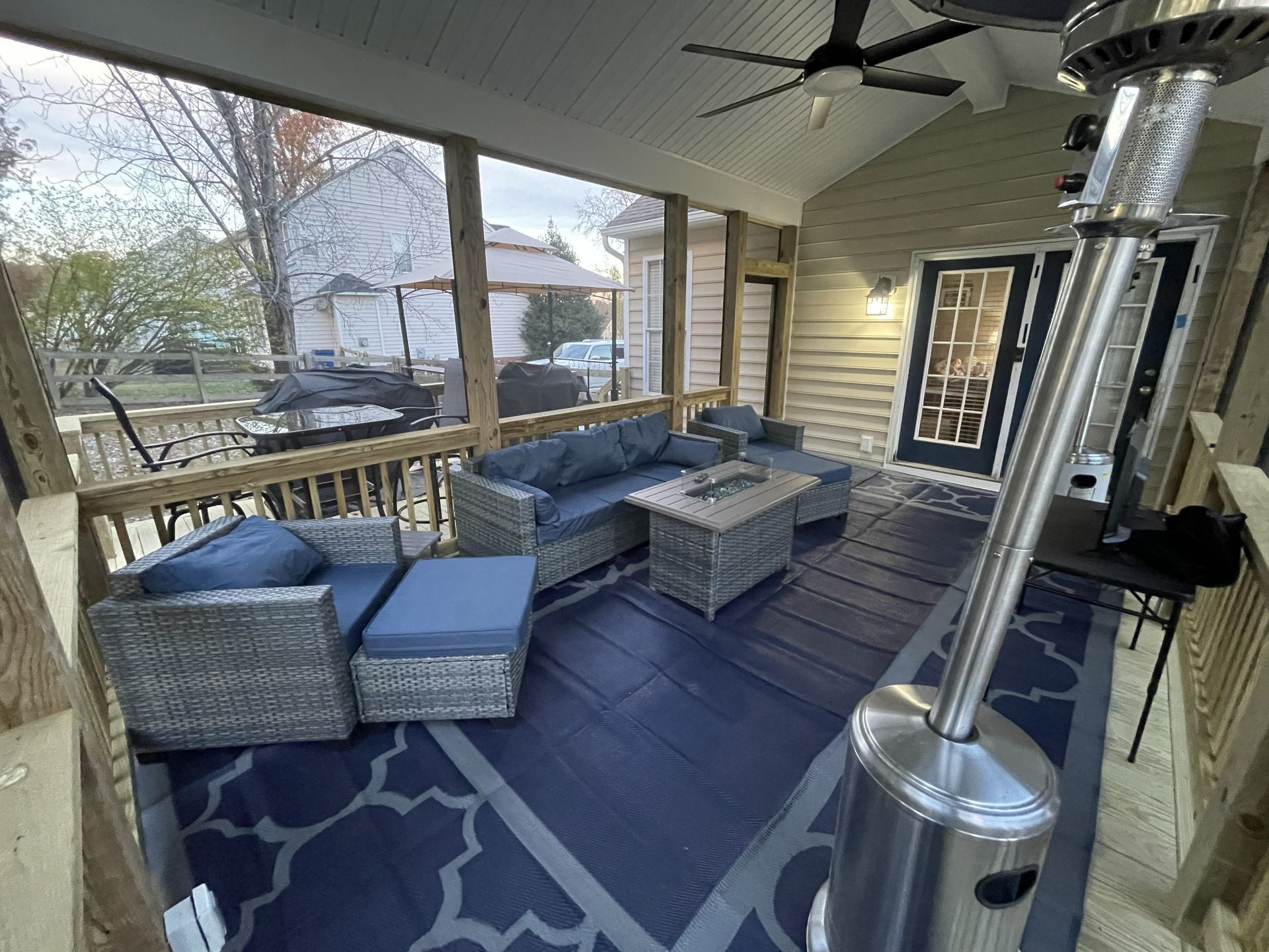 Covered patio with gray wicker seating, blue cushions, rug, and heater beside a house.