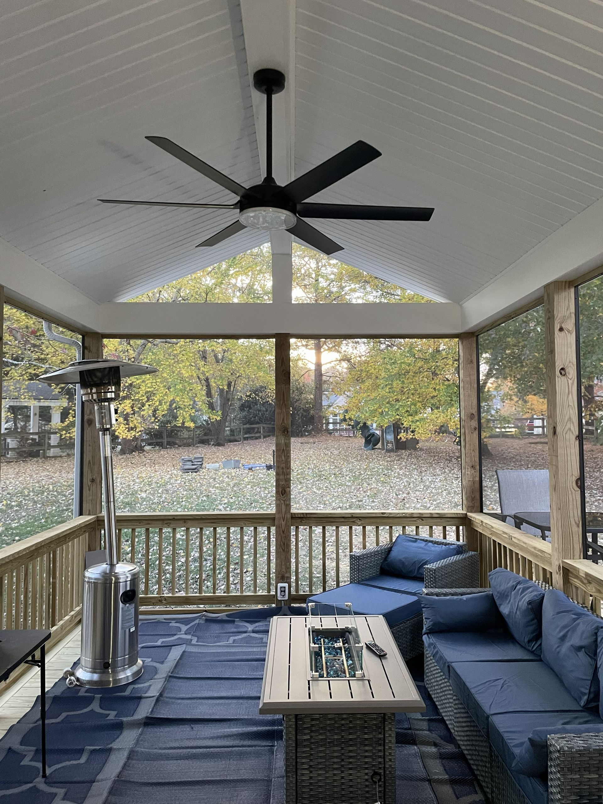Covered porch with navy sectional, coffee table, and ceiling fan overlooking a wooded yard.