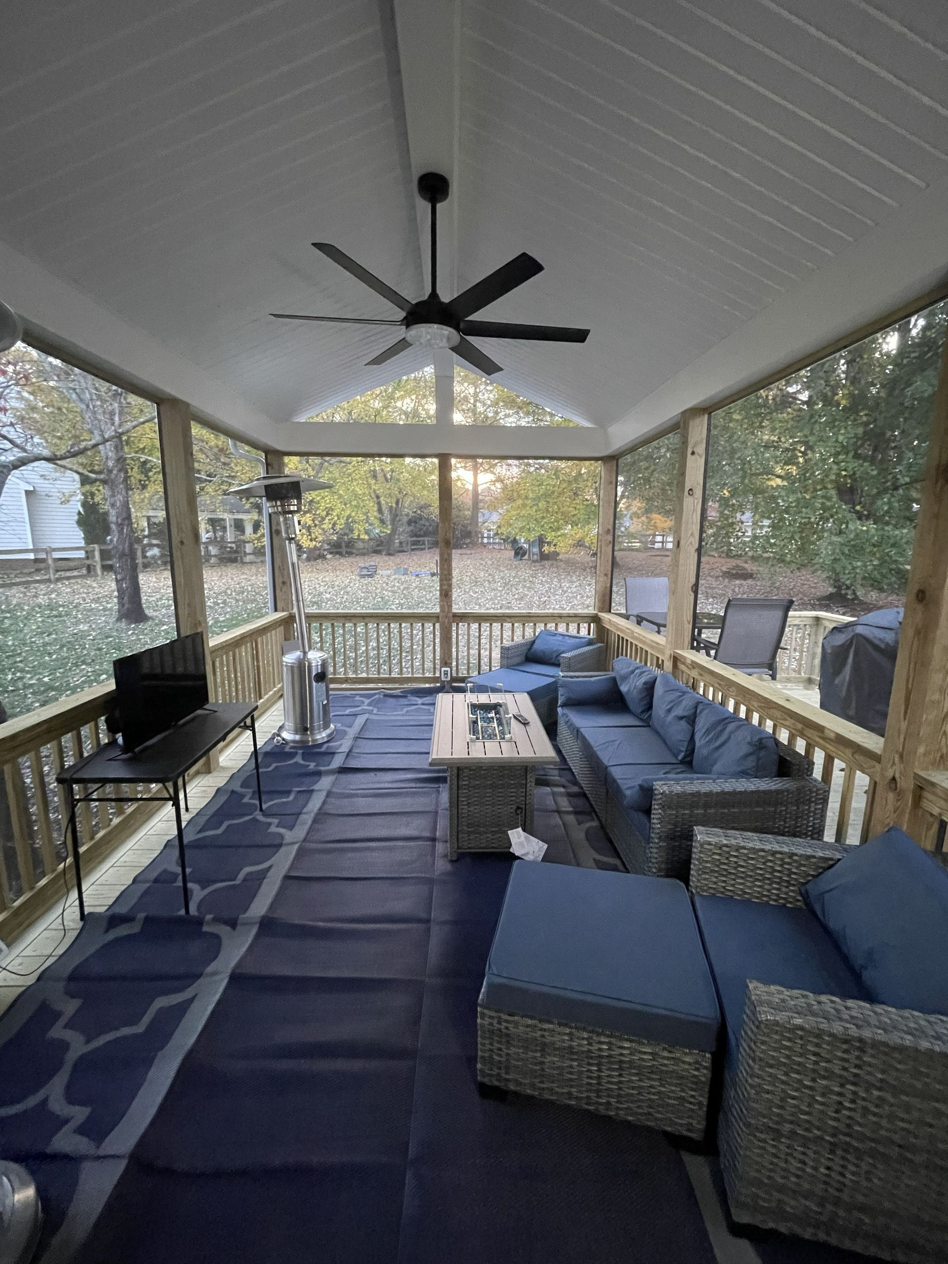 Covered outdoor patio with wicker seating, blue cushions, ceiling fan, and wooded view beyond glass railing.