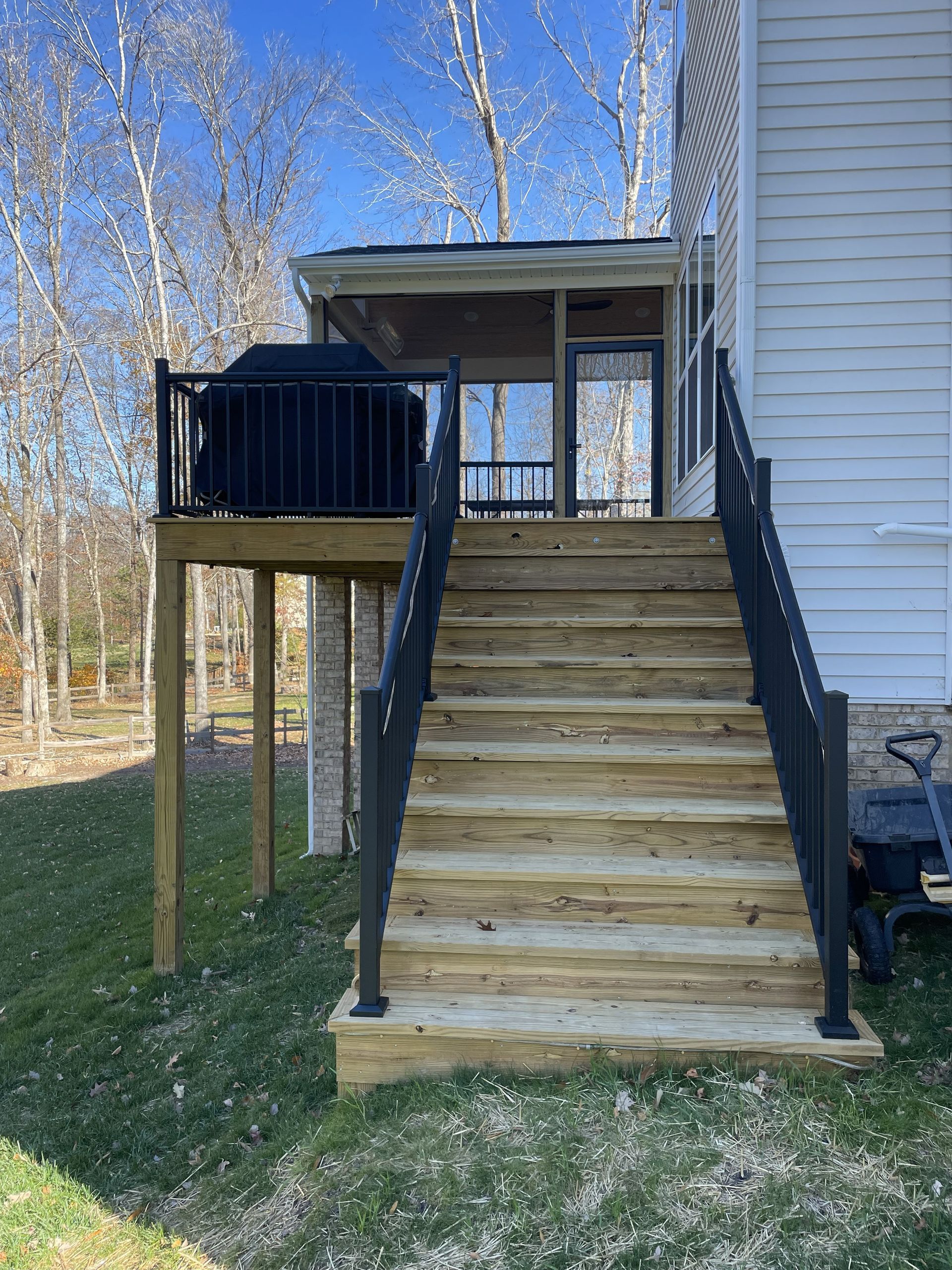 Wooden outdoor stairs with black railings leading to a raised deck beside a house, with trees in the background