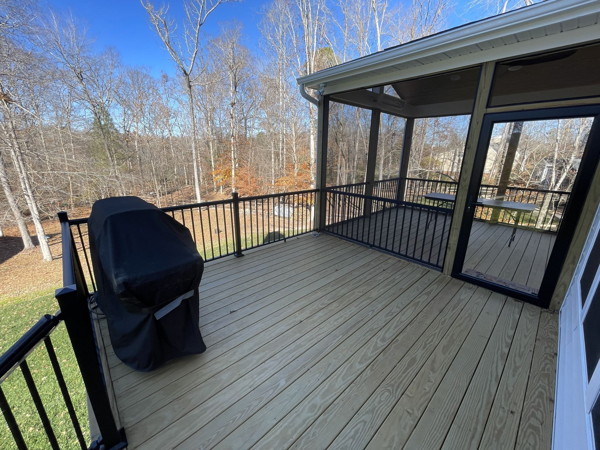 Deck with black grill beside screened porch and railing, overlooking leafless trees.