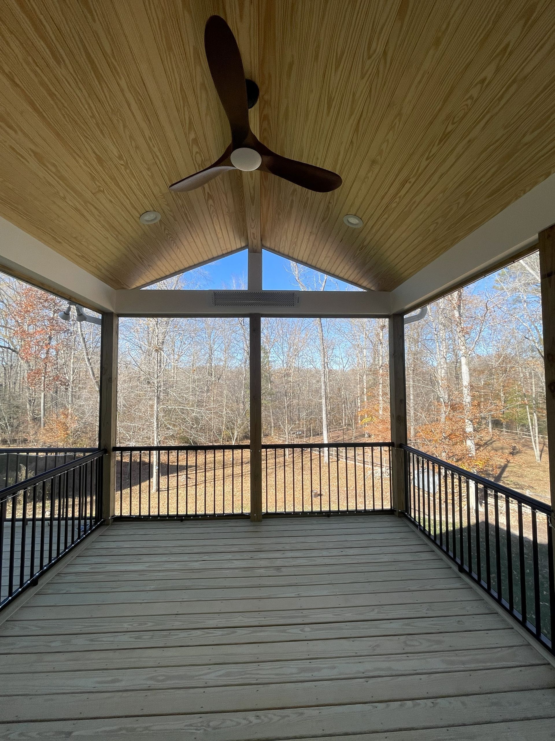 Screened porch with ceiling fan, black railing, and wooden deck overlooking autumn trees