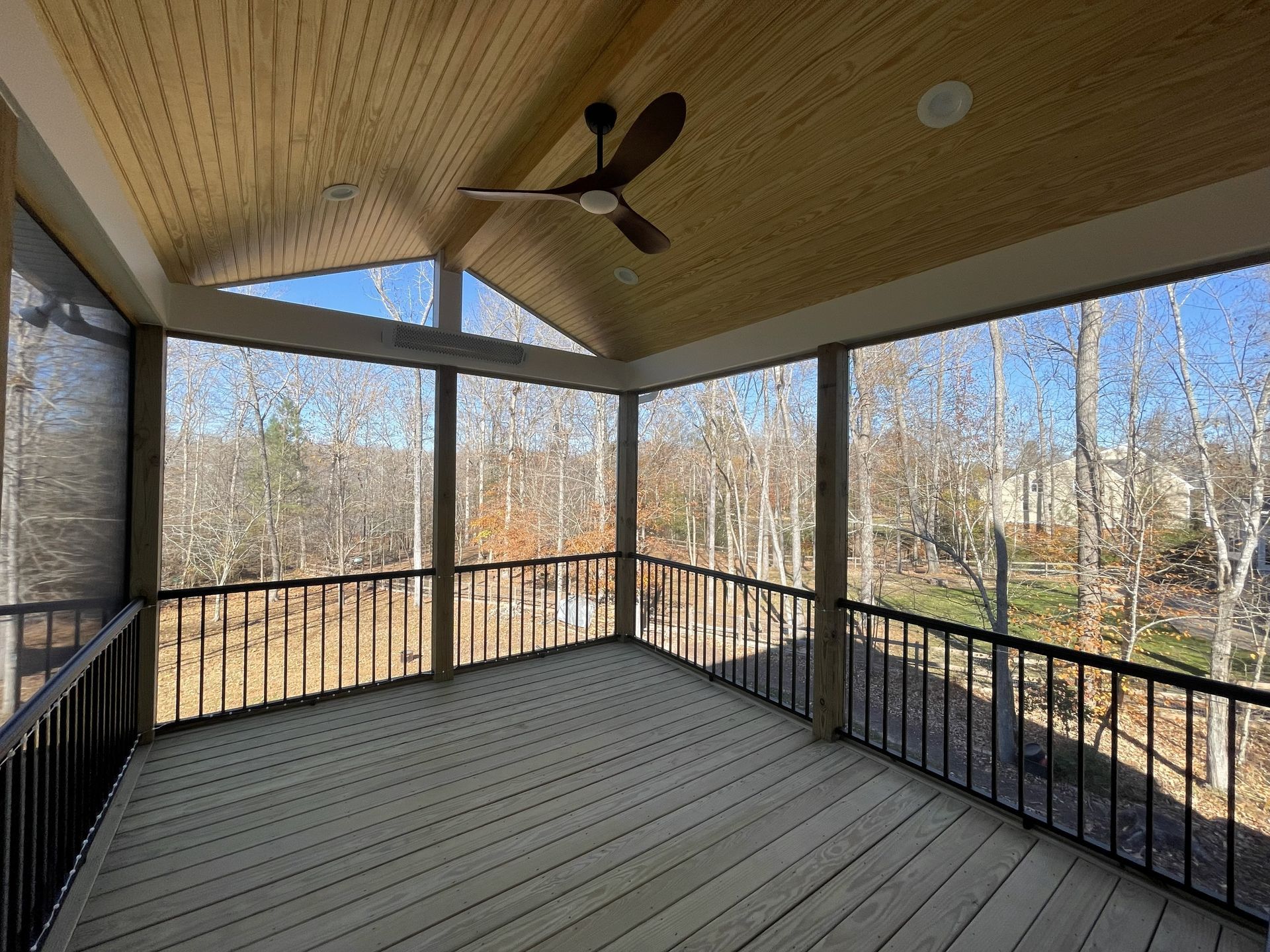 Covered screened porch with wood plank floor, black railing, ceiling fan, and forest view through mesh screens