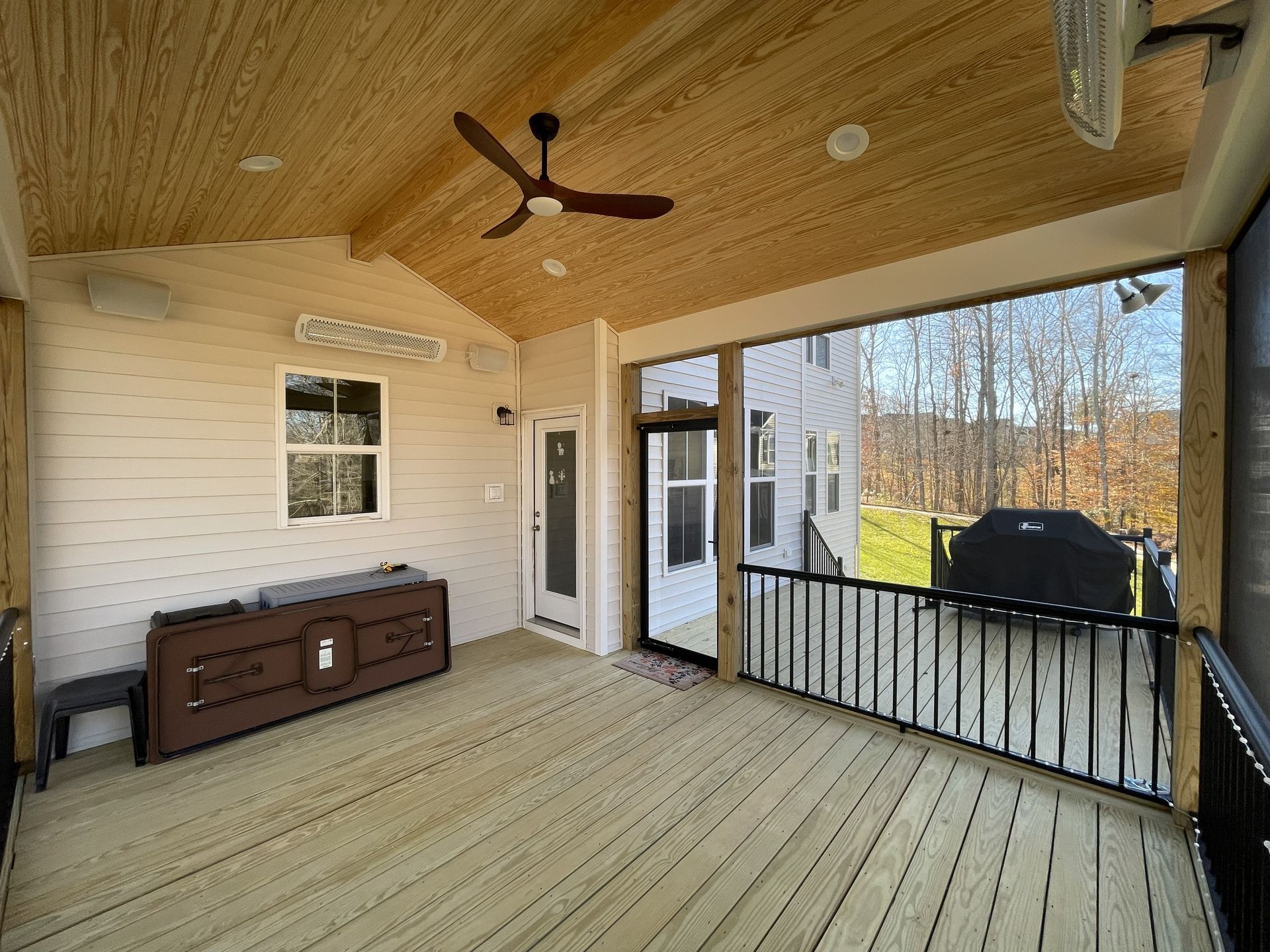 Covered wooden porch with ceiling fan, railing, bench, and view of trees and a black grill outside.