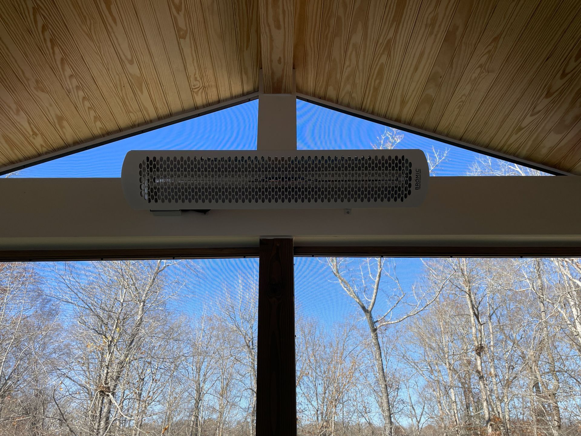 Wooden pavilion roof with a long black heater mounted above large windows, trees and blue sky outside