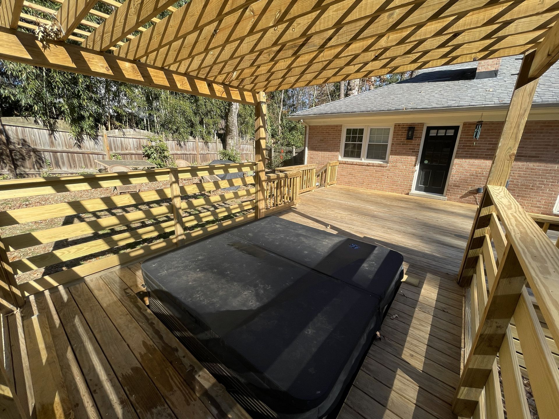 Wooden deck with a shaded pergola over a hot tub, beside a brick house and railing.