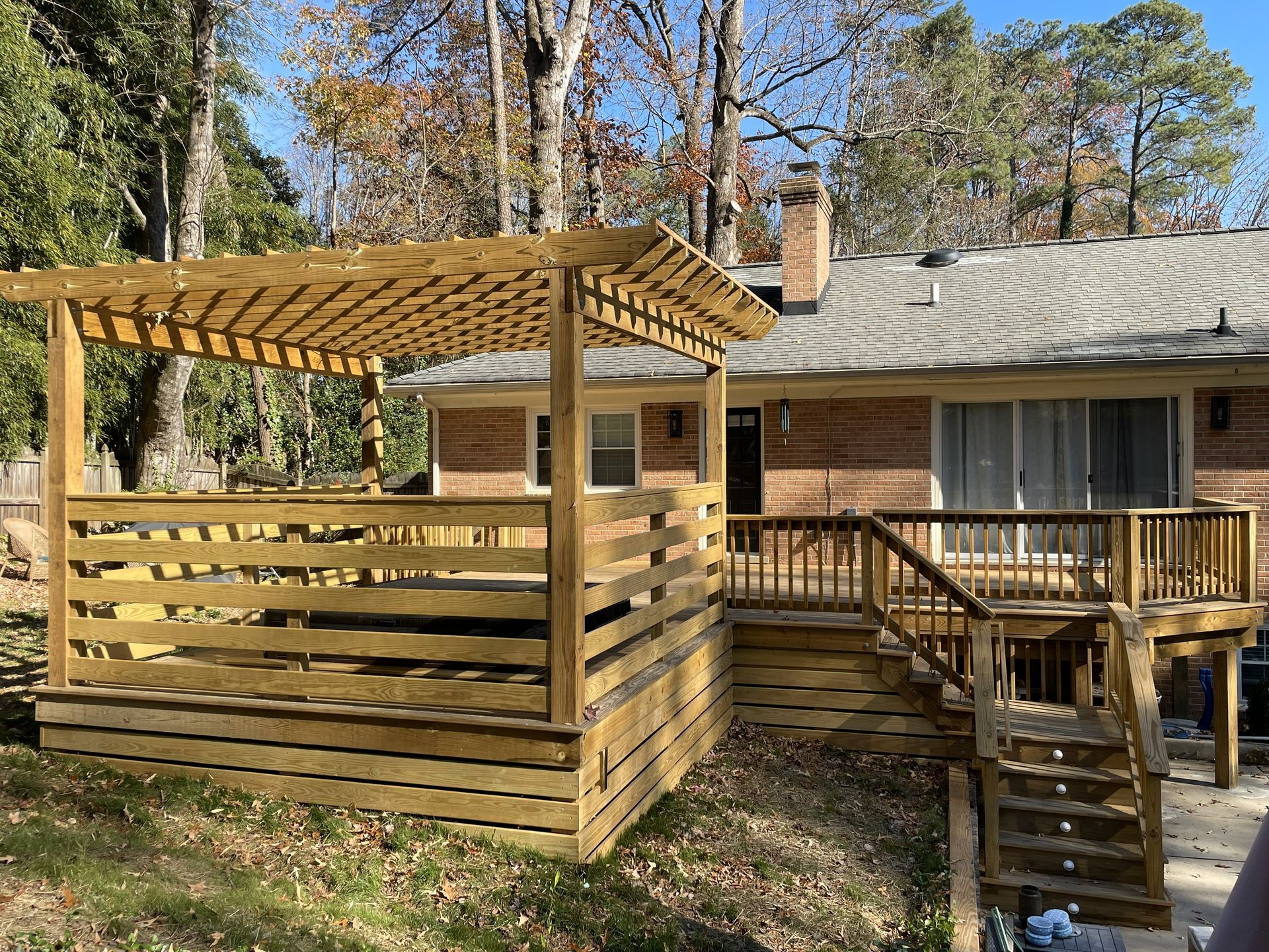 Wooden deck with pergola and railing beside a house in a wooded yard, with steps leading down