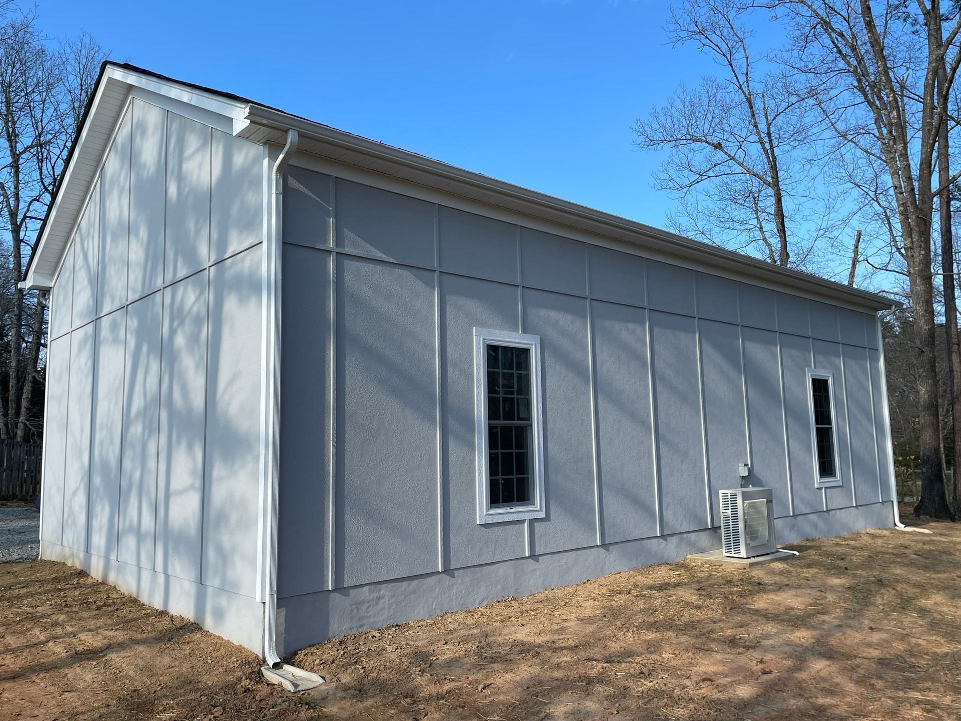Small gray building with white trim, narrow windows, and an air conditioner unit beside a wooded area.