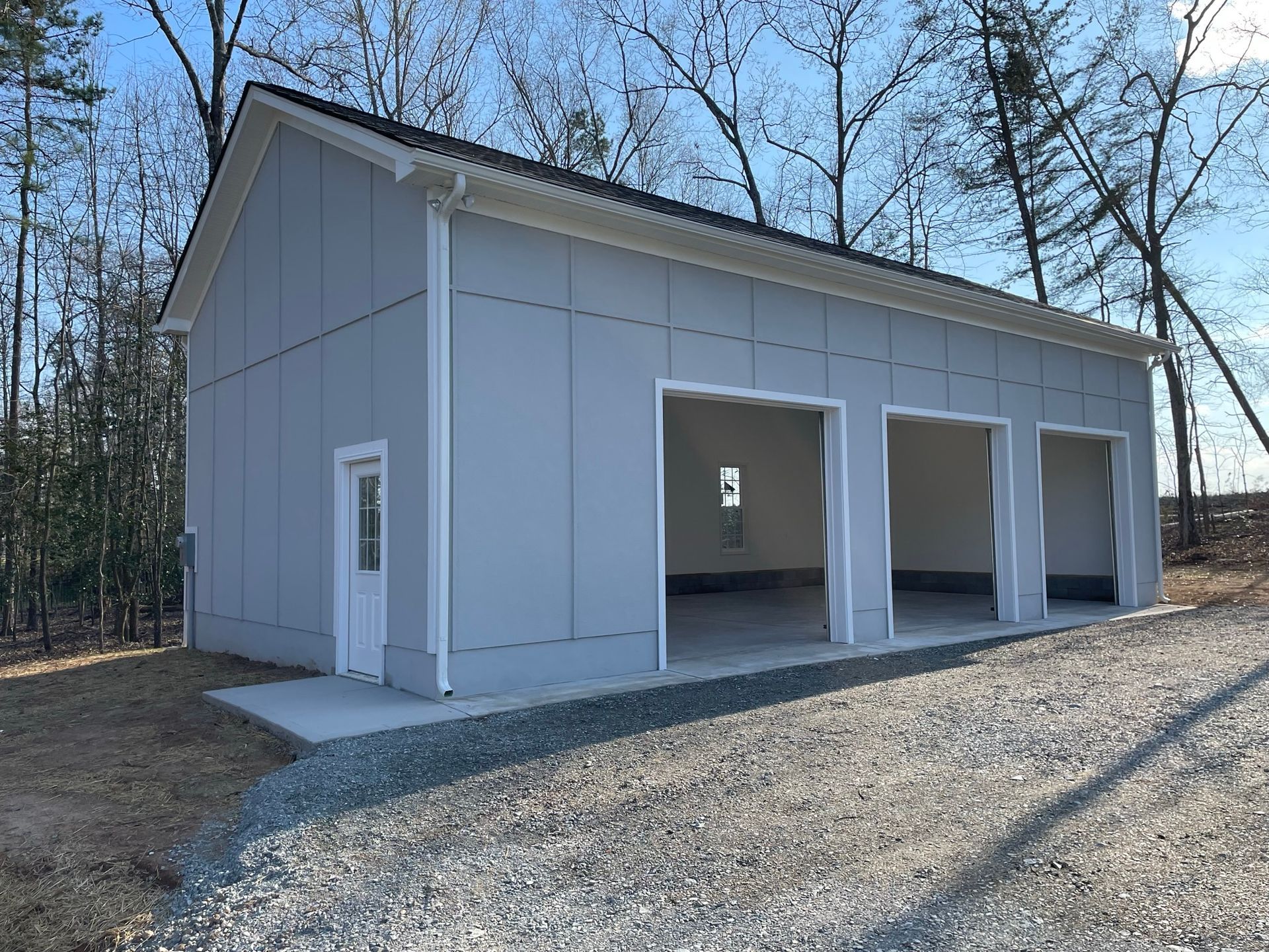 Gray metal garage building with three open bays and a side door on a gravel lot beside trees