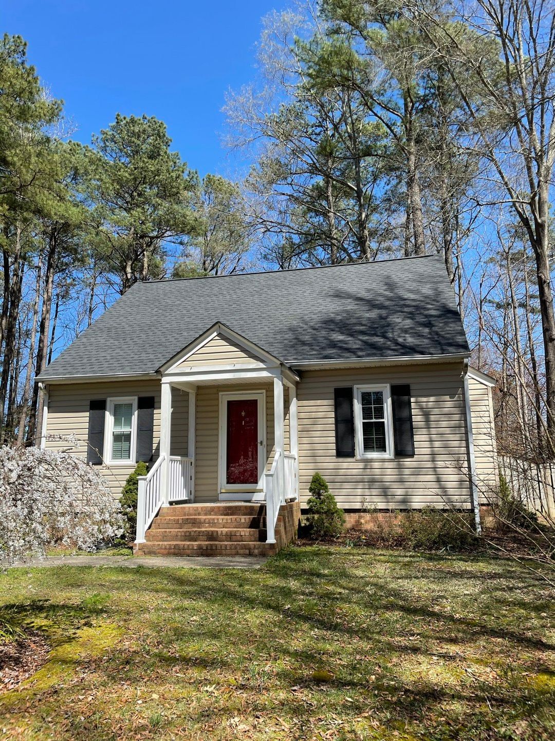 Small beige house with red door, stone front, and a front porch surrounded by trees.