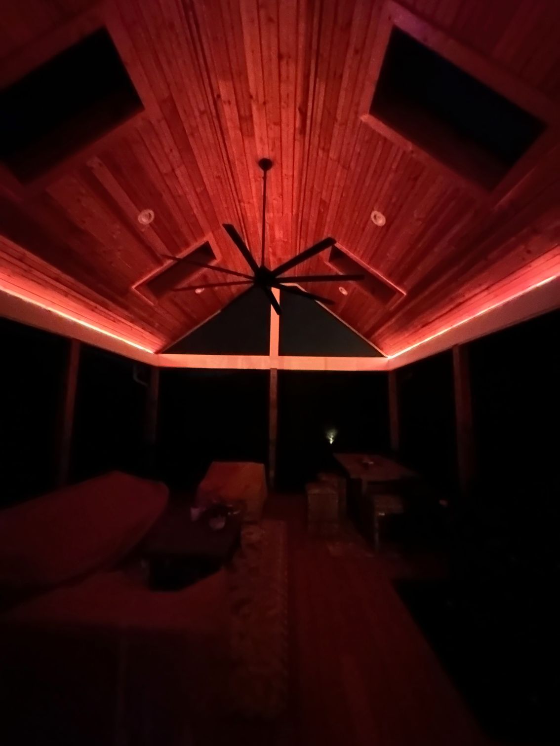 Red-lit wooden patio ceiling with central fan and lounge chairs in a dark outdoor space